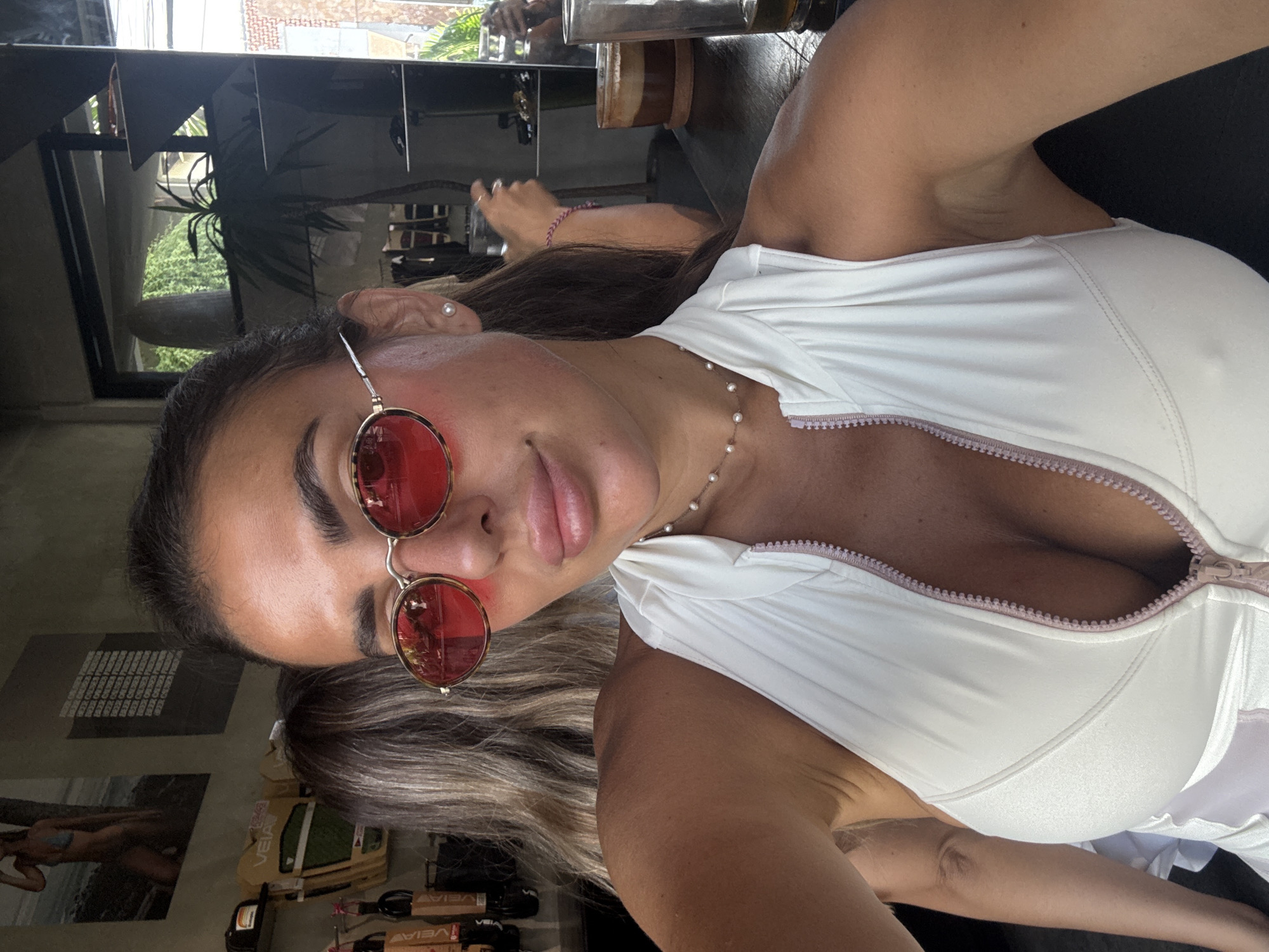 The image shows a woman wearing red sunglasses, a white top with a zipper, and a pearl necklace. She is indoors, with a window showing trees and a building in the background. There are objects such as posters and boxes visible behind her, suggesting a retail or commercial space. The location is identified as North Kuta, Indonesia.