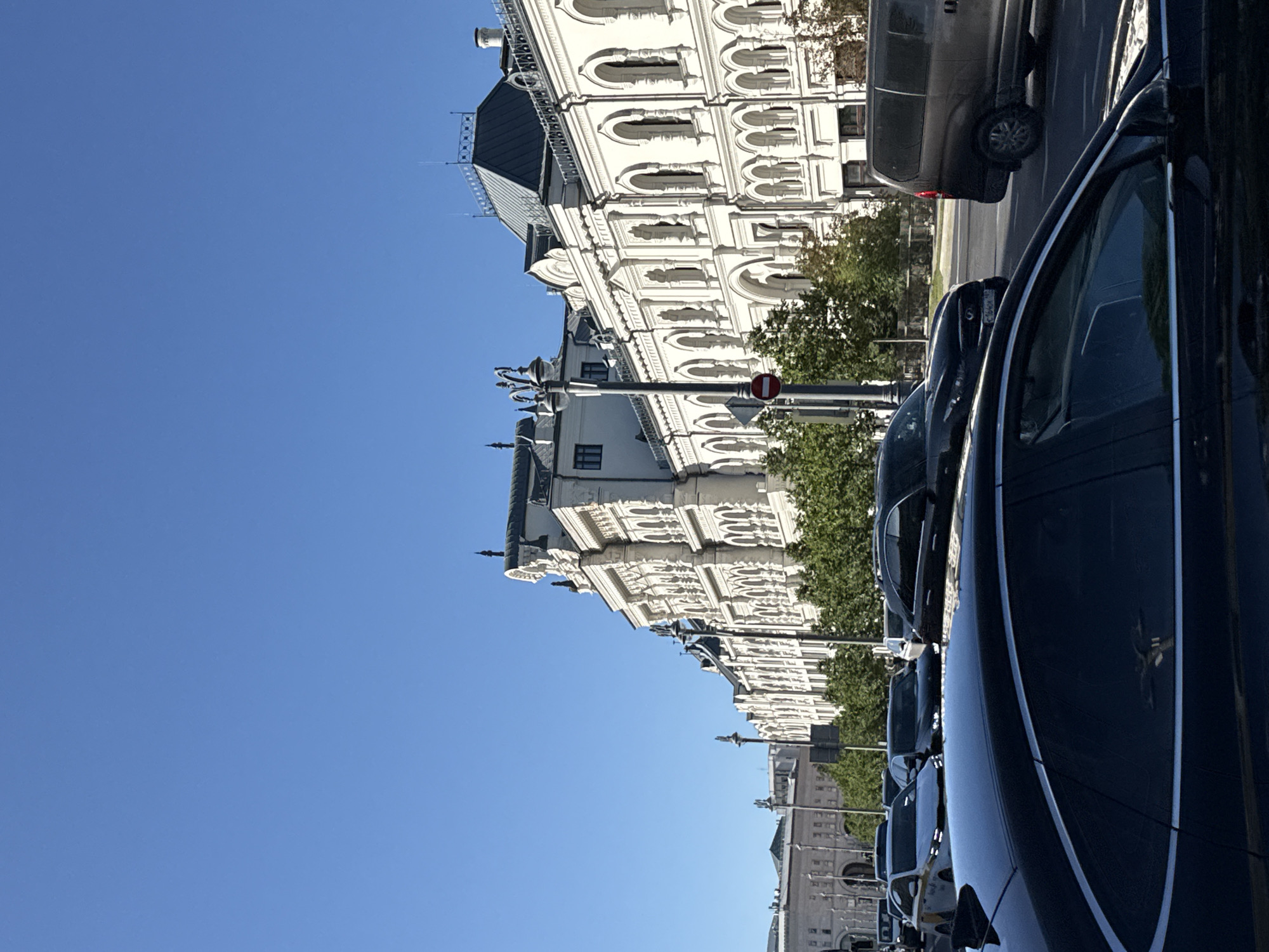 The image shows a street scene with a large, ornate white building in the background. The building has a classical architectural style with many arched windows and decorative elements. A tree with green foliage is visible in front of the building. Several cars are parked along the street, some of which are dark-colored and appear to be sedans or SUVs. The sky is a clear, bright blue, suggesting it is daytime and the weather is sunny. The perspective is slightly tilted, making it difficult to ascertain the exact orientation or precise details of the scene. There are no people clearly visible in the image. The overall impression is a view of a city street on a pleasant day.