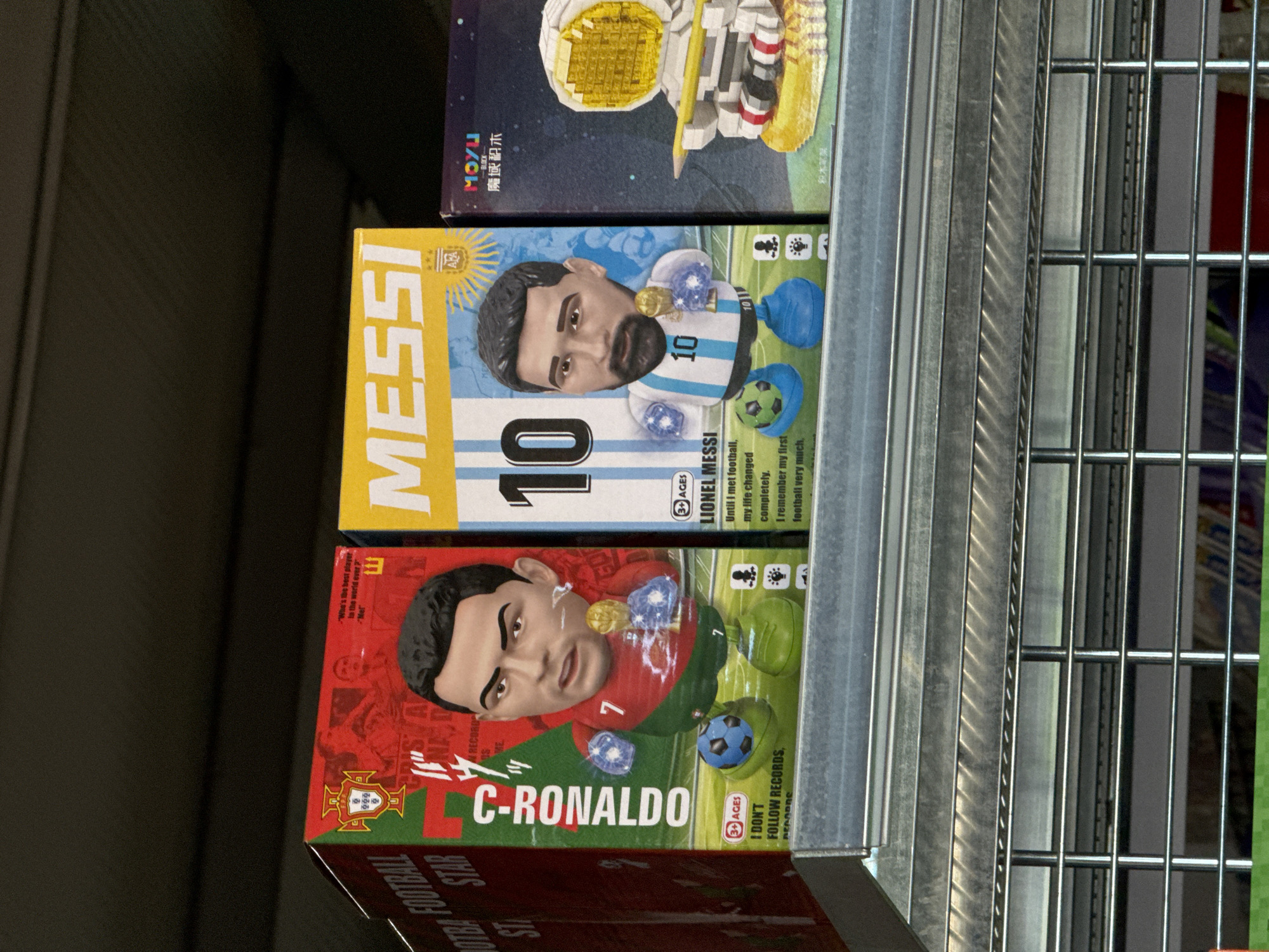The image shows a shelf in a store, likely a toy store or a novelty shop, displaying several boxes of collectible figures. On the left, a box for "C-RONALDO FOOTBALL STAR" is visible, featuring a figurine of Cristiano Ronaldo. To its right is a box for "MESSI" (Lionel Messi), also with a figurine. Further to the right, a box with a "MOYU BLOCK" logo and what appears to be a construction toy set of an astronaut is present. The store appears to be located in the city of Tsarevo, Bulgaria, as indicated by the text on the image and the surrounding context. The items are arranged on a metal shelf, with other merchandise visible in the background, suggesting a retail environment. The lighting is artificial, typical of an indoor setting. No people are visible in the frame.