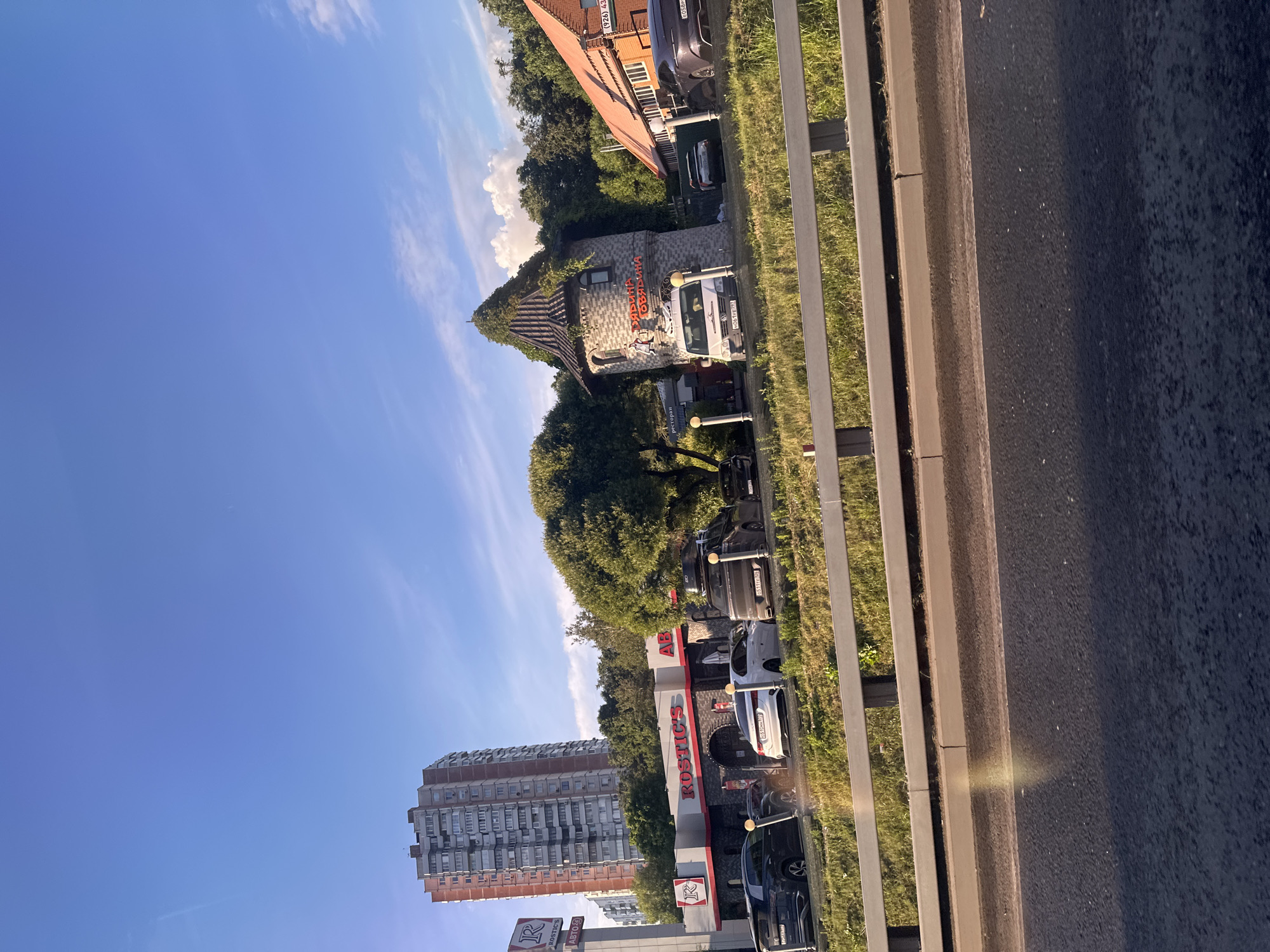 The image displays a row of buildings and cars along a roadside. The sky is blue with some wispy clouds, suggesting it is daytime, possibly late afternoon due to the low angle of the sun casting a bright reflection on the road. The scene appears to be in an urban or suburban area, with a mix of architectural styles.
One building has a distinctive, turret-like structure with a thatched roof, covered in greenery. Above the entrance, text in red illuminated letters reads "РИВЬЕРА" and "ОБЕДИМ". A white van is parked in front of this building. Adjacent to it, another building with a red-tiled roof has windows and more trees. Further down the road, a modern building with a red and grey facade and prominent signage that says "RUSTIC'S" is visible, with several cars parked in front. There's also a tall residential building with many windows in the background.
The foreground shows a paved road, a concrete curb, and a grassy area separated by a metal railing. The overall impression is a commercial or dining area situated beside a main road. There are no people clearly visible in the image. The weather appears clear and sunny.