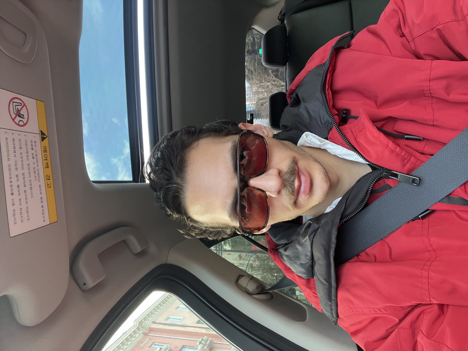 A male individual with dark, wavy hair and a prominent mustache is depicted in a selfie taken from the front passenger seat of a vehicle. He is wearing dark, rectangular sunglasses, a red puffer jacket with a black interior collar, and a white collared shirt partially visible beneath the jacket. A black seatbelt is fastened across his chest and left shoulder. The car's interior features a sunroof, through which a clear blue sky with some white clouds is visible. A white warning sticker with Korean text and a "no reclining" symbol is affixed to the passenger-side sun visor. Through the left side window, an exterior view reveals a multi-story building with light-colored, possibly beige or pale orange, facades and numerous windows, characteristic of older European-style architecture. Through the rear window, sparse trees with bare branches and distant urban structures are observable. The scene is located on a street in Tbilisi, Georgia, identifiable by the distinctive architectural style of the visible buildings.