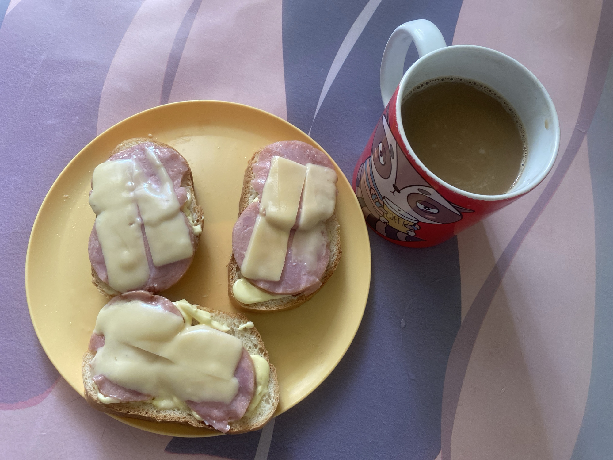 The image displays a meal set on a patterned surface, likely a placemat or tablecloth, in an indoor setting in Chaikovsky, Russia. The main subjects are three open-faced sandwiches on a yellow plate and a red mug filled with a beverage.
The sandwiches consist of slices of white bread topped with a creamy spread (possibly mayonnaise), several slices of pink meat (appearing to be ham or sausage), and melted light yellow cheese. On two of the sandwiches, the cheese is cut into strips, while on the third, it appears to be a single, larger melted slice. The cheese has a slightly glossy, melted appearance.
Beside the plate, to the right, sits a red ceramic mug with a white handle. The mug features a cartoon illustration of a raccoon-like animal. Partially visible text on the mug appears to say "COFF," suggesting "COFFEE." The mug is filled with a light brown liquid, consistent with coffee or tea, possibly with milk or cream. Small bubbles are visible on the surface of the drink.
The background surface beneath the plate and mug has a wavy, abstract pattern in shades of purple, pink, grey, and light blue, creating a gentle gradient effect. The lighting suggests daytime. No people are visible, and no specific activity is taking place other than the presentation of the meal.