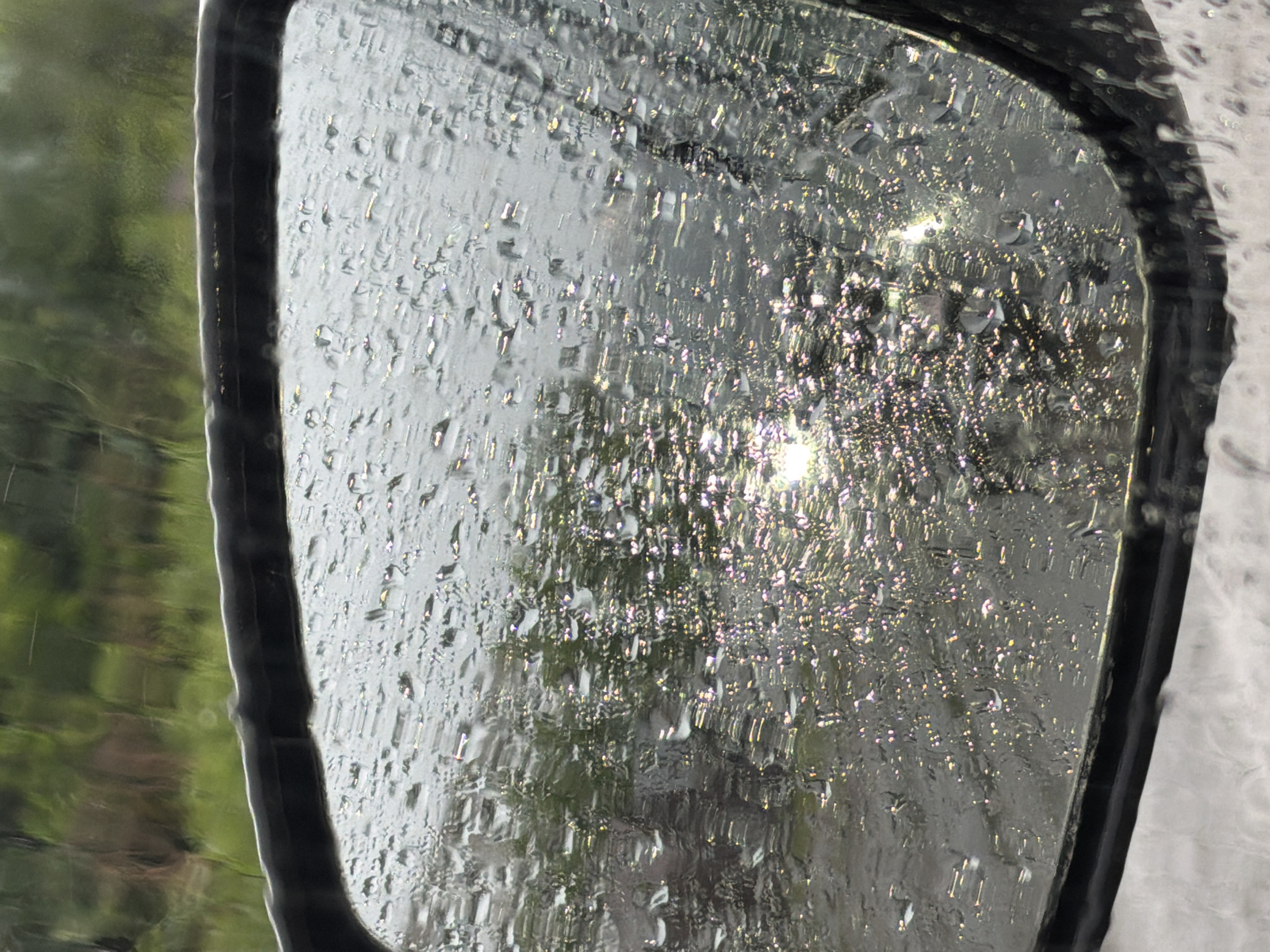 The image shows a close-up of a car's side-view mirror covered in raindrops. The mirror reflects an outdoor scene, possibly a road or landscape with green foliage and the sun's reflection creating a sparkling effect. The rain distorts the reflection, with water droplets creating a textured and abstract appearance on the mirror's surface. The surrounding environment appears overcast or rainy. There are no people or specific landmarks visible that would indicate a location in Chaikovsky, Russia.