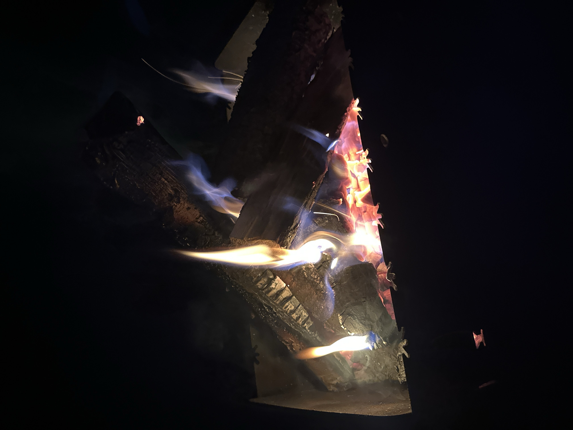 The image shows a close-up view of a wood fire burning inside a metal fire pit or stove. Several logs are arranged in a pyramid-like structure, with flames rising upwards. The flames are primarily orange and red, with some blue and white areas indicating higher temperatures. Smoke is visible, swirling around the burning logs. The background is dark, suggesting the photo was taken at night or in a dimly lit environment. There is no evidence to suggest the location is Biysk, Russia. There are no people in the image.
