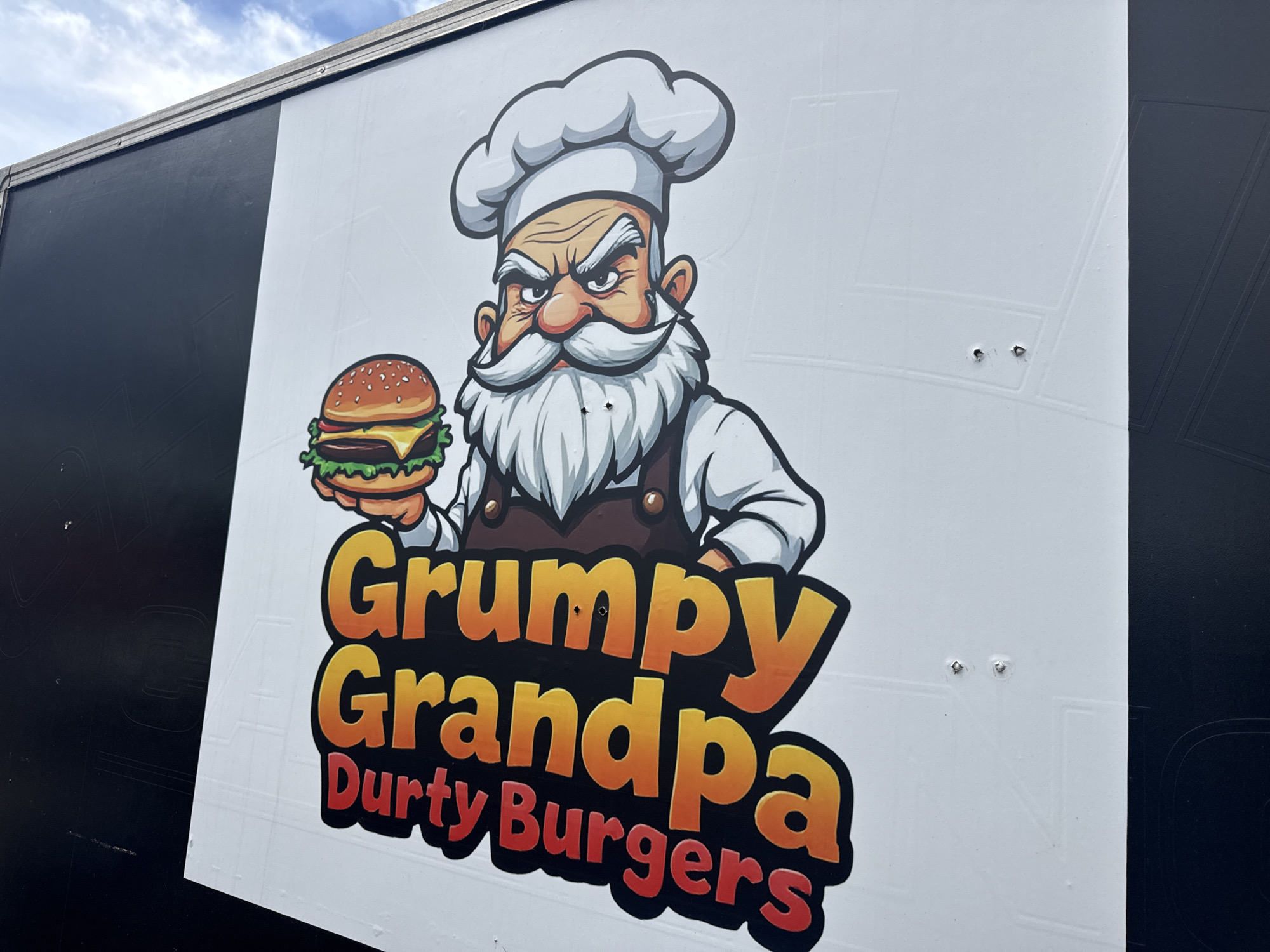 The image shows a white trailer with a large graphic on its side. The graphic features a cartoon of a grumpy-looking chef, identified by the text as "Grumpy Grandpa Durty Burgers." The chef is holding a burger. The trailer is parked outdoors, under a cloudy blue sky. The overall scene appears to be in an outdoor setting, possibly a public space or a business lot. There are no people visible in the image. The trailer is primarily black and white, with the graphic dominating the white section. The graphic itself is colorful, with the text rendered in bold, gradient lettering. The image is taken from a slightly low angle, looking up at the trailer.
