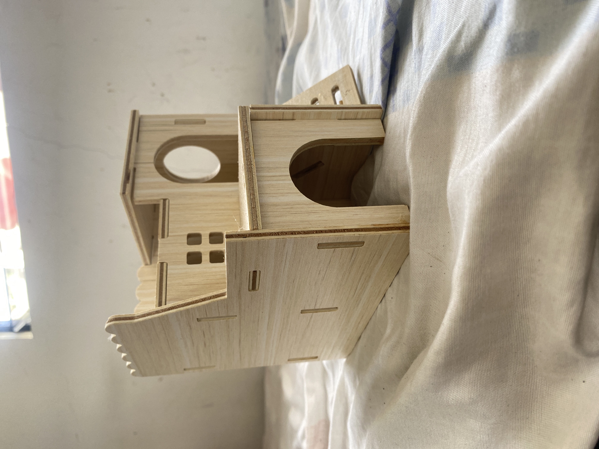A light-colored, unfinished wooden multi-level hamster playhouse sits on a textured fabric surface. The playhouse has a castle-like facade with crenellations along the top edge of one side. It features several openings, including square windows and a circular opening on an upper level, as well as an arched entrance on a lower level. A ramp extends from one side of the playhouse. The fabric beneath the playhouse appears to be a light-colored cloth with subtle blue patterns, possibly a blanket or tablecloth. The background shows a blurred white wall with some discoloration and a window with light filtering through. The overall setting suggests an indoor environment. There are no people visible in the image. The image does not contain any information or context to suggest a location in Davao City, Philippines.