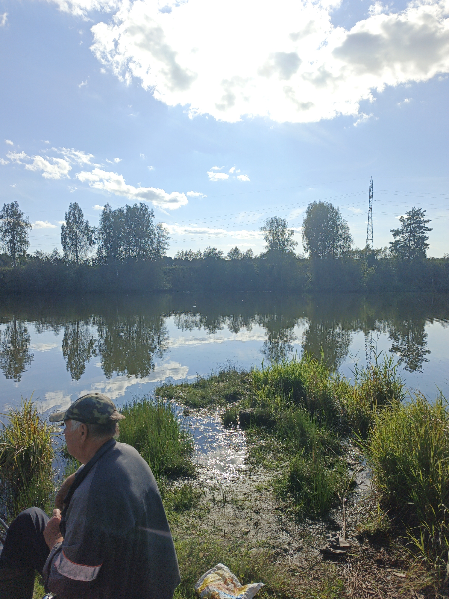 The image shows a tranquil waterside scene with a person fishing. The sky is a bright blue with scattered white clouds, and the sun is shining, casting reflections on the water. The water's edge is lined with lush green grass and reeds. In the background, a dense line of trees separates the water from a clear blue sky. A power line tower stands tall against the horizon. The person, wearing a camouflage baseball cap and a dark shirt with reflective stripes on the sleeves, is seated on the bank, facing away from the camera and towards the water, holding a fishing rod. There is a fishing reel and other fishing gear visible near the person. The overall impression is one of a peaceful, natural setting ideal for fishing.