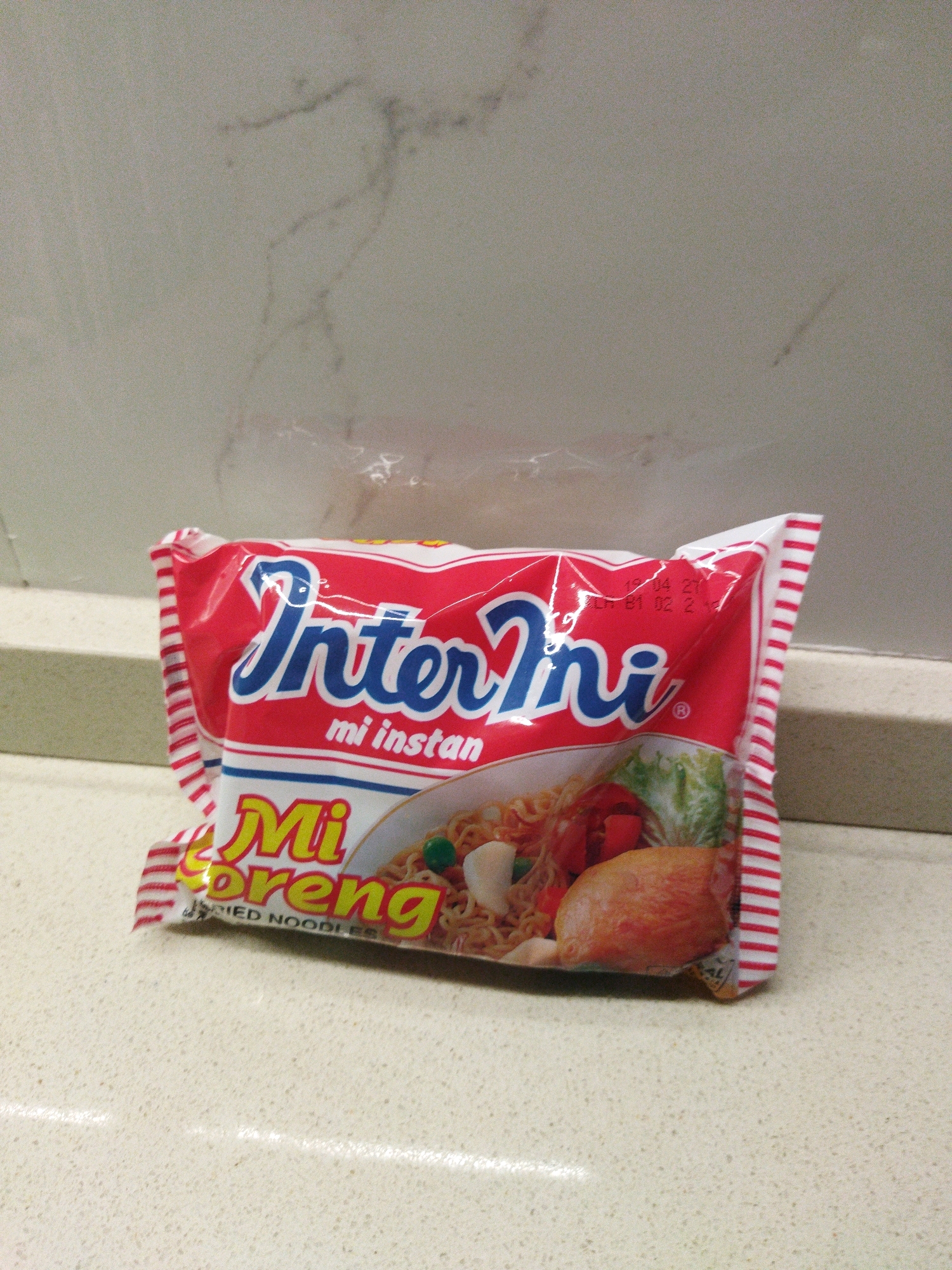 A single packet of Indomie Mi Goreng instant noodles is positioned centrally on a speckled, light-colored countertop. The packet is primarily red with white and blue lettering. The brand name "Indomie" is prominently displayed in blue cursive script across the upper half of the packet. Below this, the text "mi instan" appears in smaller white lettering. The lower portion of the packet features the words "Mi Goreng" in yellow, bold lettering. The image on the packet depicts a serving of noodles with various toppings, including what appears to be a piece of chicken or meat, vegetables, and a hard-boiled egg. The packet also has a red and white striped border along the left and right edges. The background consists of a light-colored wall with subtle gray markings. The overall scene is indoors, likely in a domestic or commercial setting, with no indication of its specific location within Banjar Agung, Indonesia, other than the general context of the product.