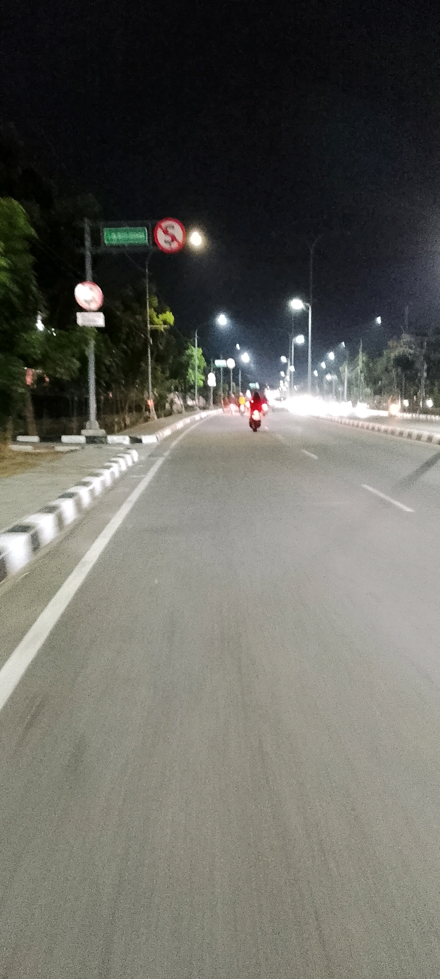 The image depicts a road at night in Gunung Putri, Indonesia. A motorcycle with a rider in a red jacket is visible in the middle of the road, heading away from the viewer. Streetlights illuminate the scene, casting a glow on the asphalt road and the surrounding trees. Traffic signs, including a "no U-turn" sign, are present on the left side of the road. The road is bordered by a sidewalk with black and white striped markings. The overall atmosphere is that of a quiet, potentially suburban, road at night.