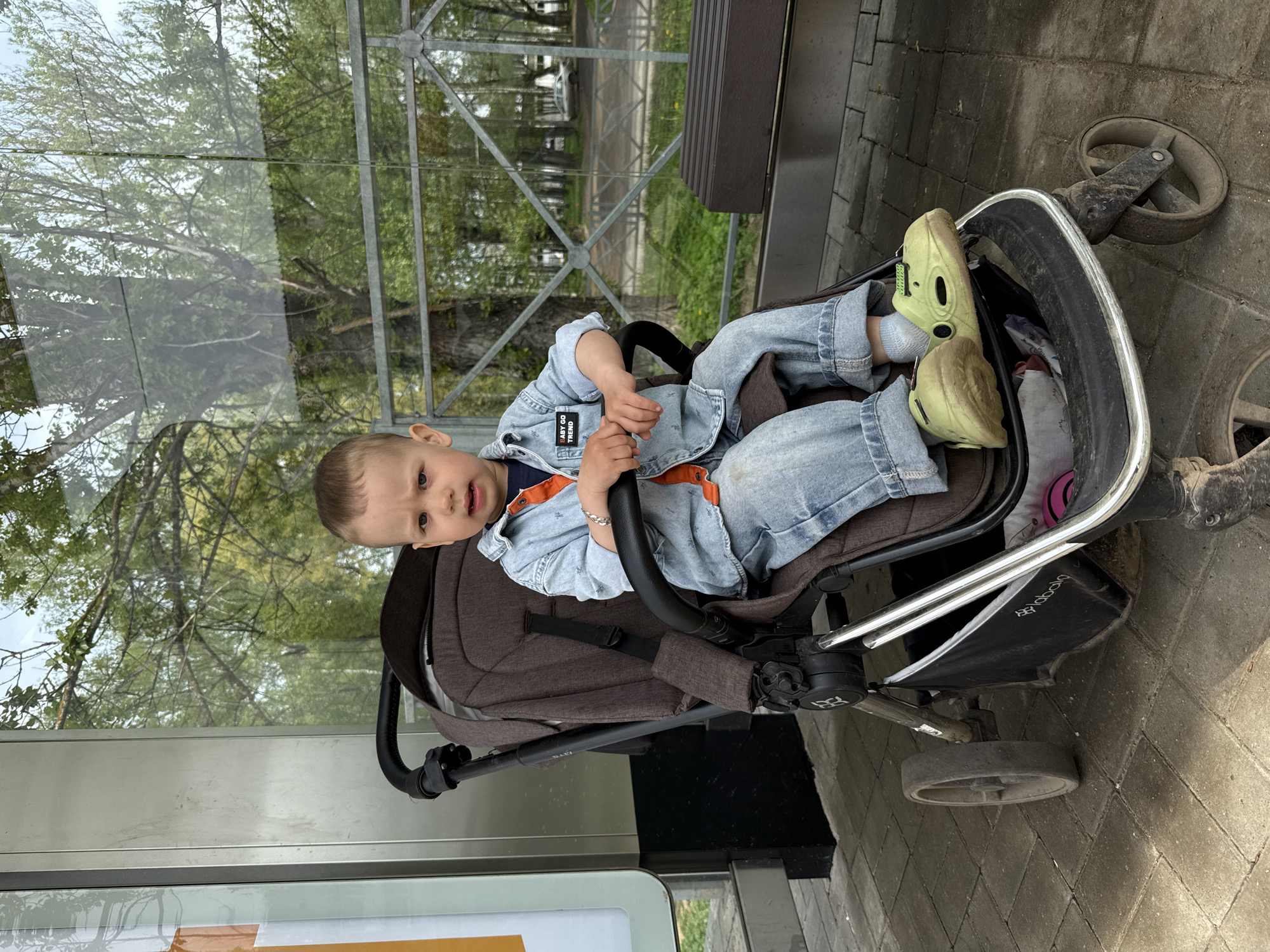 A young child is sitting in a stroller next to a bus shelter. The child is wearing a denim jacket, blue jeans, and yellow sandals. The stroller is facing the street, and the bus shelter is behind it. The city of Rusanovo, Russia, is visible in the background.