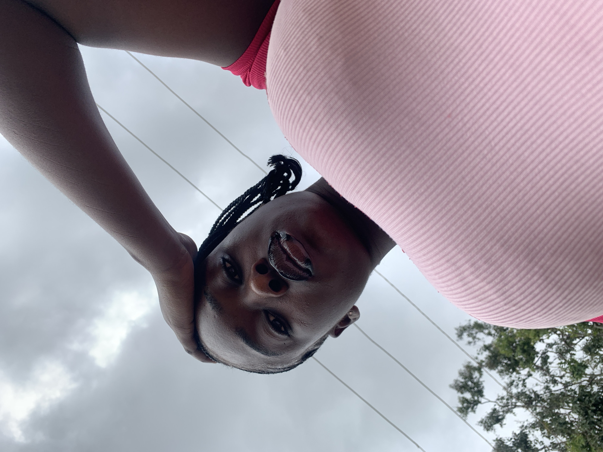 A low-angle, close-up, outdoor shot captures a young person with dark skin, wearing a pink ribbed top and a red garment underneath. Their left hand is raised, touching the top of their head, and their hair is styled in braids. The person's lips are dark and glossy, and their eyes are looking forward. The background consists of a cloudy sky, overhead power lines, and the tops of green trees, suggesting a location in Nabatum, Nigeria. The lighting is overcast, creating a diffused effect.