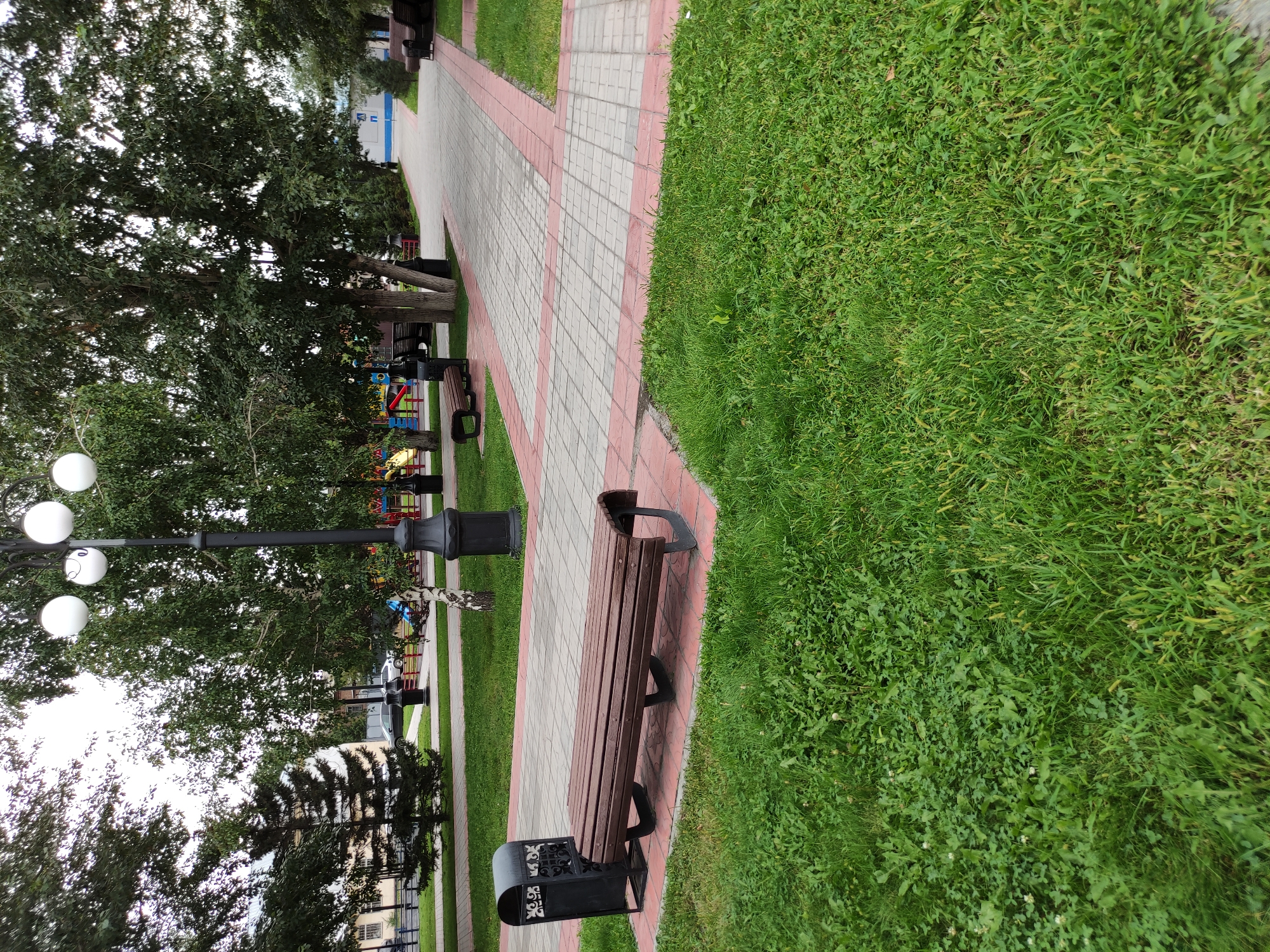 A paved walkway with red and white square tiles winds through a grassy park area in Semey, Kazakhstan. A dark brown wooden bench with black metal legs sits on a small bricked area beside a dark gray trash receptacle with decorative metalwork. Beyond the bench, a lamppost with four white globes stands tall, surrounded by trees with lush green leaves. In the background, a playground with colorful structures is visible, along with more benches and trees. The sky above is overcast, suggesting a cloudy day.