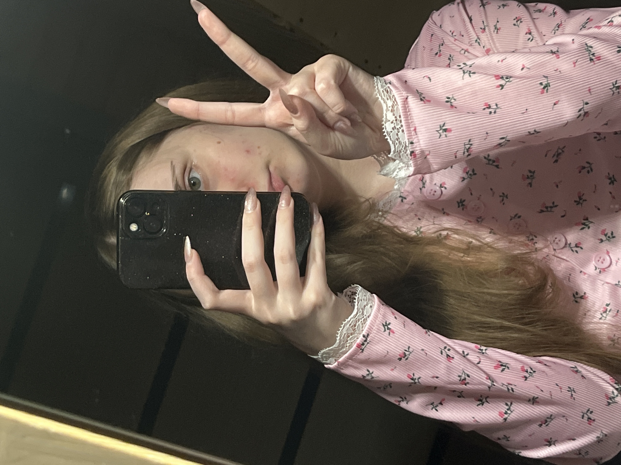 A young woman with blonde hair, wearing a pink floral button-up shirt with lace trim, takes a mirror selfie. She holds a black smartphone in her left hand, partially obscuring her face, and makes a peace sign with her right hand. The background is dark and indistinct, suggesting an indoor setting. There is no information provided about the location in the city of Ashkadar, Russia, or any environmental details beyond the interior setting.