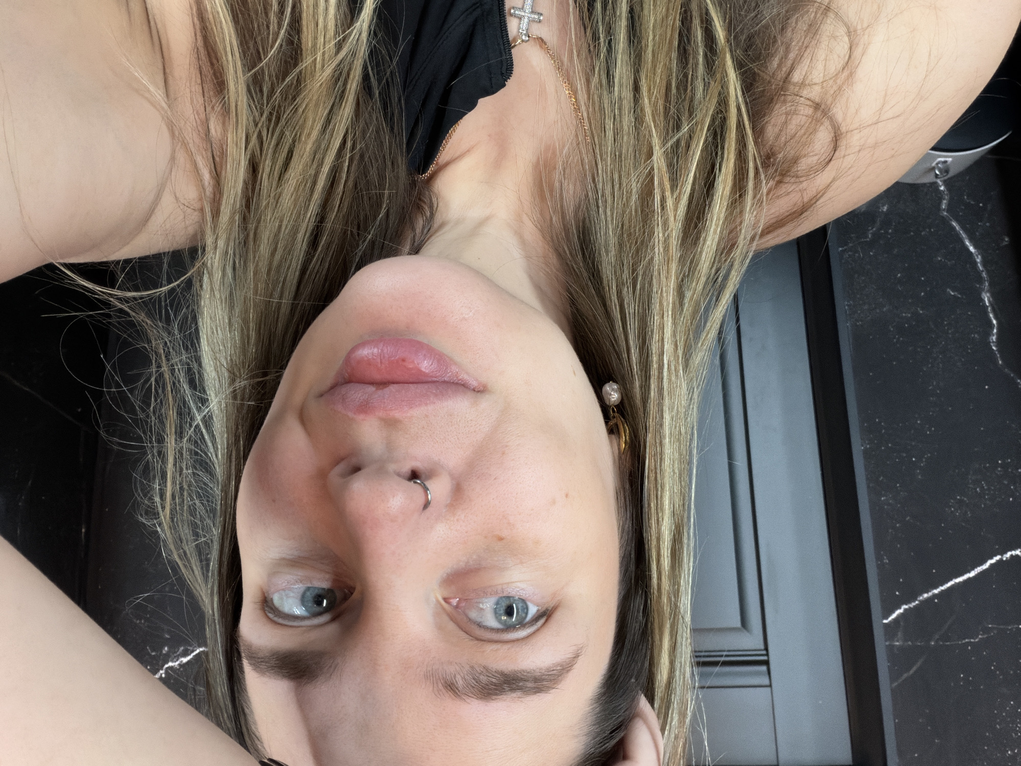 A fair-skinned woman with light brown and blonde hair is positioned upside down in the frame. Her eyes are blue, and she has a nose ring on her left nostril. She is wearing a black top, with a necklace featuring a cross pendant visible. She also has an earring on her right ear. The background includes a dark grey cabinet or door, next to a countertop with a black marble effect. The location is not specified, and there are no interactions or actions taking place.