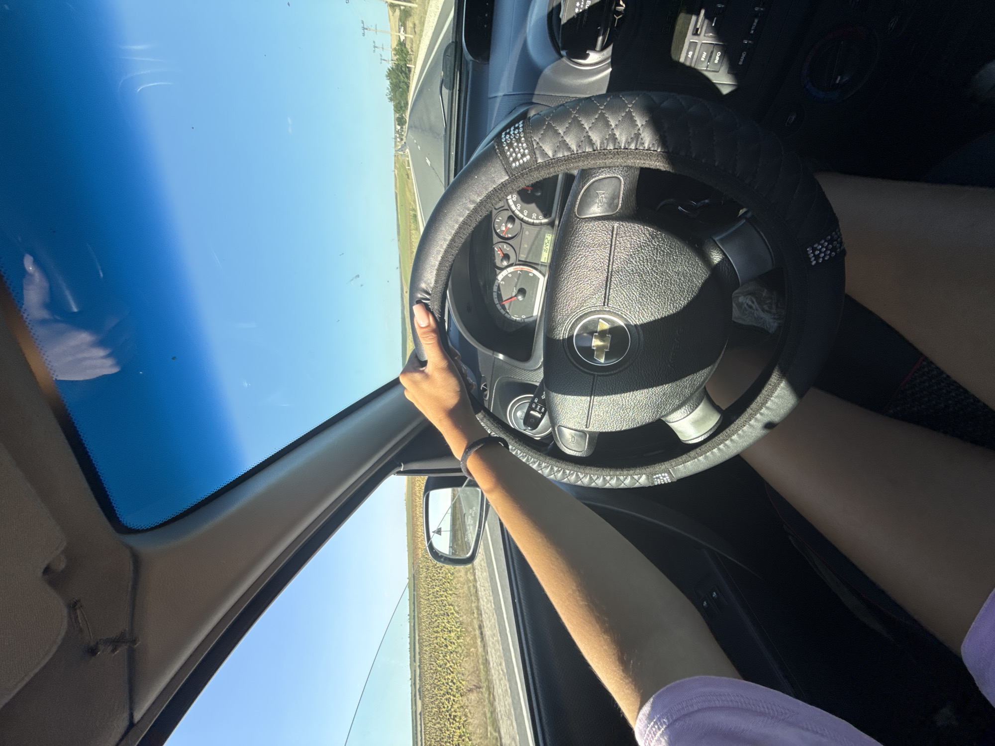 A first-person perspective shot shows a person's hands on the steering wheel of a car, driving down a road. The car's interior is visible, including the dashboard with gauges and a stereo system. Outside the car, a sunny day is depicted with a clear blue sky and a vast field of sunflowers stretching into the distance. The road appears to be a highway, with other vehicles visible in the distance. The scene is captured in a bright, natural light, suggesting it is daytime. The location is identified as Comrat, Moldova.