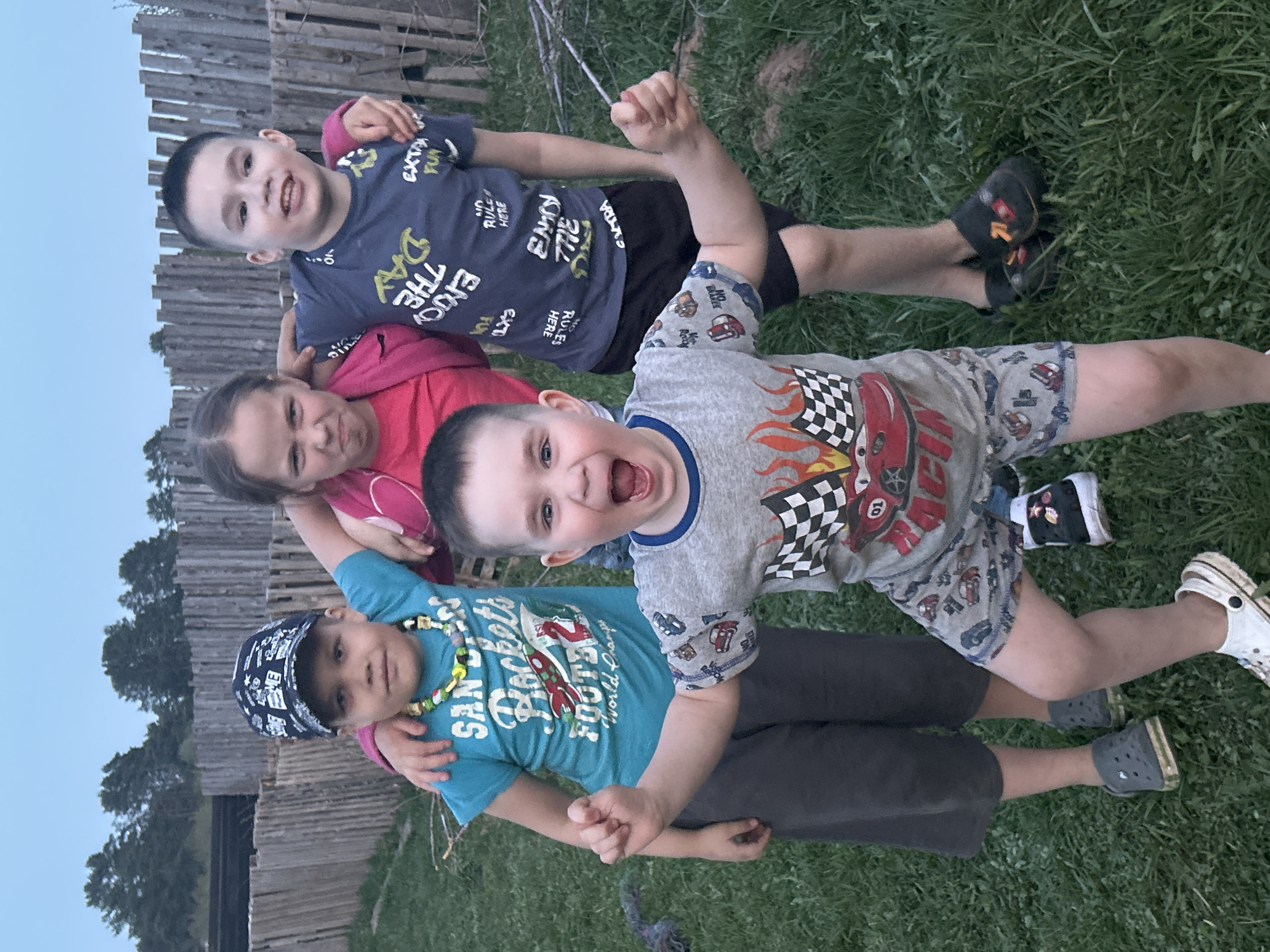 Four children lie on a grassy area, likely outdoors. The background shows a wooden fence and trees, possibly indicating a rural or suburban setting. From top to bottom: a boy with a dark blue t-shirt; a girl with a pink hoodie; a boy with a gray t-shirt; a boy with a teal t-shirt and black cap. The boy with the gray t-shirt has his mouth open, possibly yelling. All are dressed in casual clothing. No context indicates a location in the city of Rusalevka, Russia.