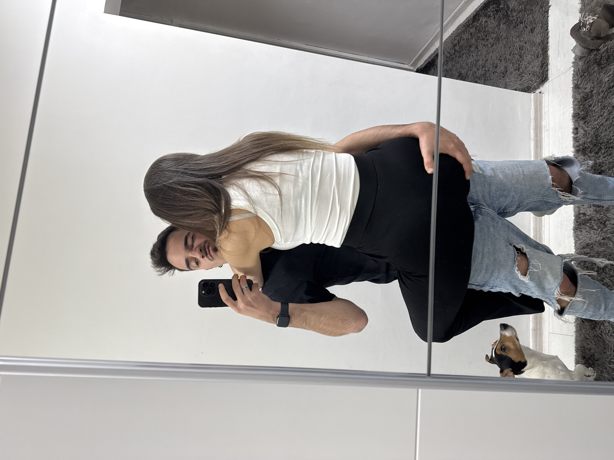 This image captures a joyful mirror selfie of a couple and their dog in what appears to be a home in Sofia, Bulgaria. The man, positioned on the left, holds a black iPhone with his right hand to take the photo, smiling warmly towards the camera. He has short dark hair, a light beard, and is wearing a black t-shirt, ripped light blue jeans, and a black smartwatch on his left wrist. A silver ring is visible on his right hand.
The woman is embracing him from behind, her face mostly obscured as she leans her head on his shoulder. She has long brown hair and is dressed in a white ribbed tank top and black leggings.
On the right side of the image, reflected on the floor, a small tri-color dog, possibly a Jack Russell Terrier, is lying playfully on its back with its paws in the air, looking upwards. The indoor setting features a large mirror reflecting a bright white wall, a light-colored wooden floor, and a shaggy gray rug. The scene is well-lit, suggesting daytime, and the expressions convey a happy, relaxed moment between the subjects. No discernible text is visible within the image.