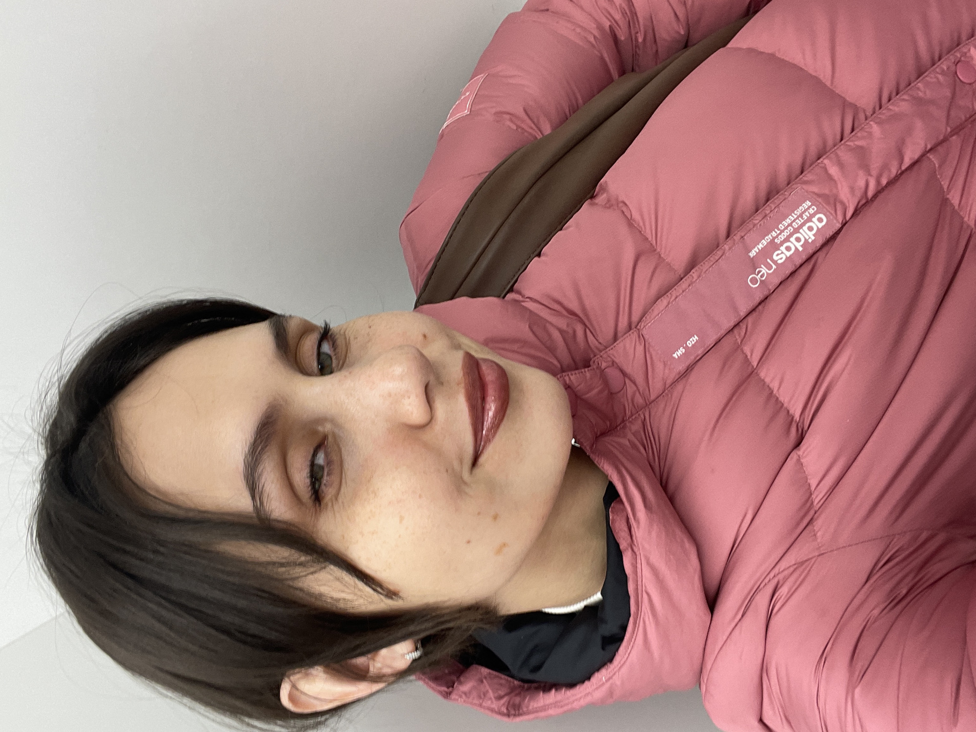The image shows a woman wearing a pink Adidas Neo puffer jacket. She has dark hair and light skin with freckles. The background is a plain white wall. There is no information provided in the image to suggest that the photo was taken in Izhevsk, Russia.