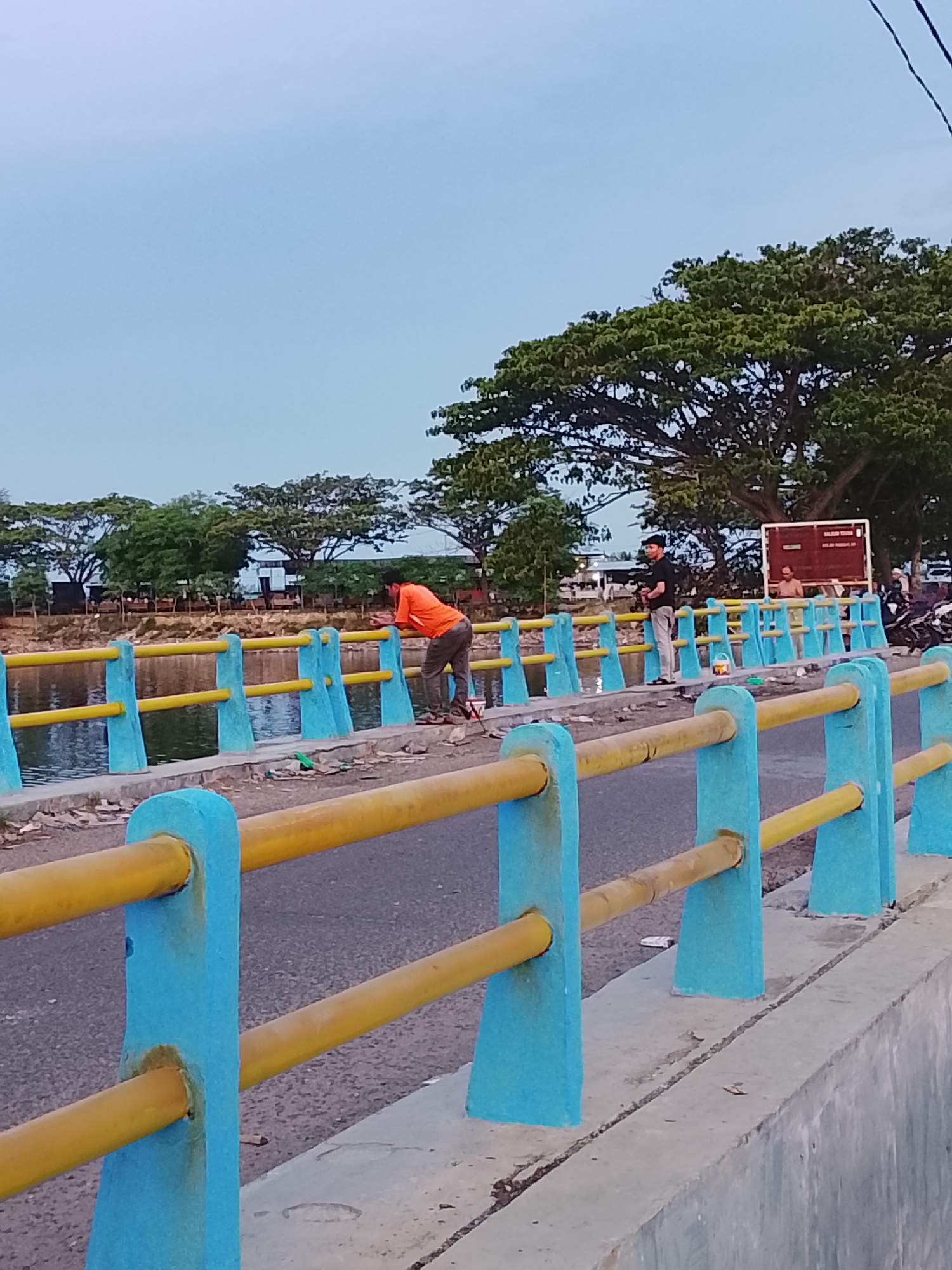 In Lhokseumawe, Indonesia, two individuals are present on a bridge. One person, wearing an orange shirt and brown pants, is leaning over the blue and yellow railing, looking down at the water. The second person, in a black shirt and grey pants, stands further along the bridge, holding what appears to be a fishing rod. The bridge has a concrete embankment on the right and a waterway below on the left. Beyond the bridge, a line of trees and some buildings are visible under a pale blue sky. A signpost with red text is also present in the background. The scene suggests a casual outdoor activity.