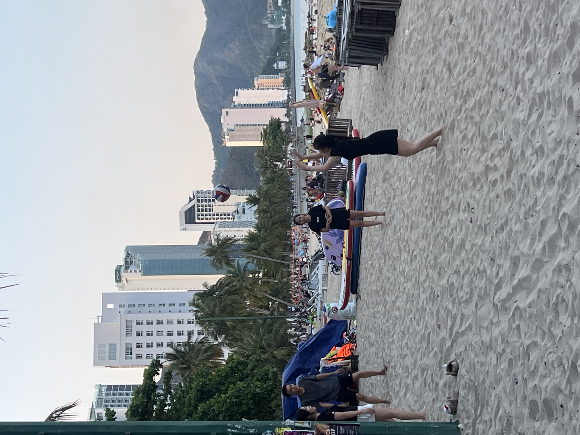 This image captures a vibrant beach scene in Bắc Nha Trang, Vietnam, likely during the late afternoon or early evening. The sandy beach is a hub of activity, with a backdrop of modern high-rise buildings and a prominent mountain range under a clear sky.
In the foreground, two women are engaged in a game of volleyball. One woman, dressed in a black top and shorts, stands near two surfboards or paddleboards, while another, wearing a black dress, is captured mid-action, reaching up to hit a white, blue, and red volleyball that is airborne. Several pairs of shoes or sandals are scattered on the sand nearby.
Further back, under a blue tarp or tent, a small group of people are seen relaxing. The mid-ground reveals numerous wooden beach chairs, some stacked and some occupied by beachgoers. Palm trees are interspersed along the beach, separating the immediate activity from the urban development.
The background showcases a bustling coastal city with many tall, contemporary buildings lining the shore. Faintly visible on one white building are the letters "HOTEL". Beyond the city, a large, rugged mountain dominates the horizon. The ocean is visible between the beach and the distant city buildings.
Overall, the scene depicts a popular recreational area where people are enjoying leisure activities by the sea, with a mix of natural beauty and urban development. On a sign in the very bottom left corner, part of the word "POP" is visible.