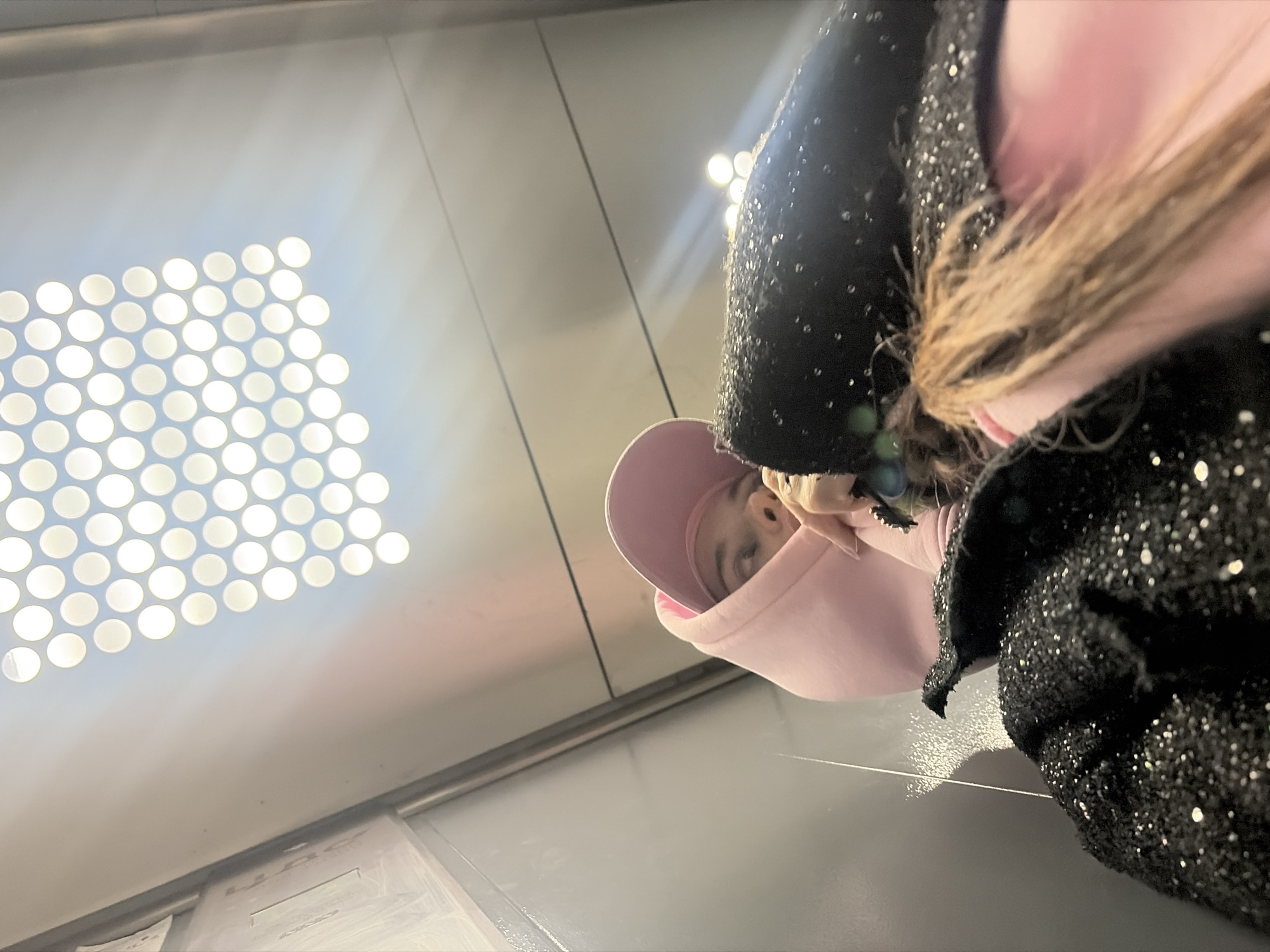 A low-angle shot shows a person wearing a pink hooded sweatshirt and a black sparkly jacket, seemingly taking a selfie in an elevator. The elevator has a grid of circular lights on the ceiling and gray walls. The location is Saint Petersburg, Russia.