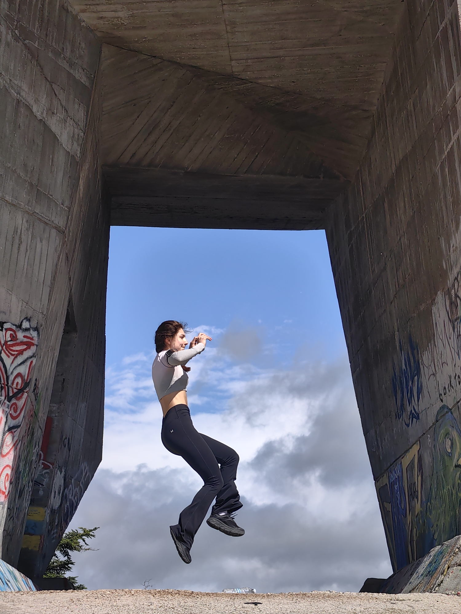 A woman is photographed leaping in mid-air against the backdrop of a concrete structure in Varna, Bulgaria. She wears a long-sleeved, light-colored crop top and dark-colored athletic pants and shoes. The concrete structure appears to be a large, unfinished building or architectural element, possibly a part of a bridge or tunnel. The concrete surface is marked with graffiti. The sky is visible through the opening of the structure, revealing a mix of blue sky and clouds. The ground below is somewhat uneven. The woman's arms are raised. The overall environment suggests an urban, possibly abandoned or under-construction site.