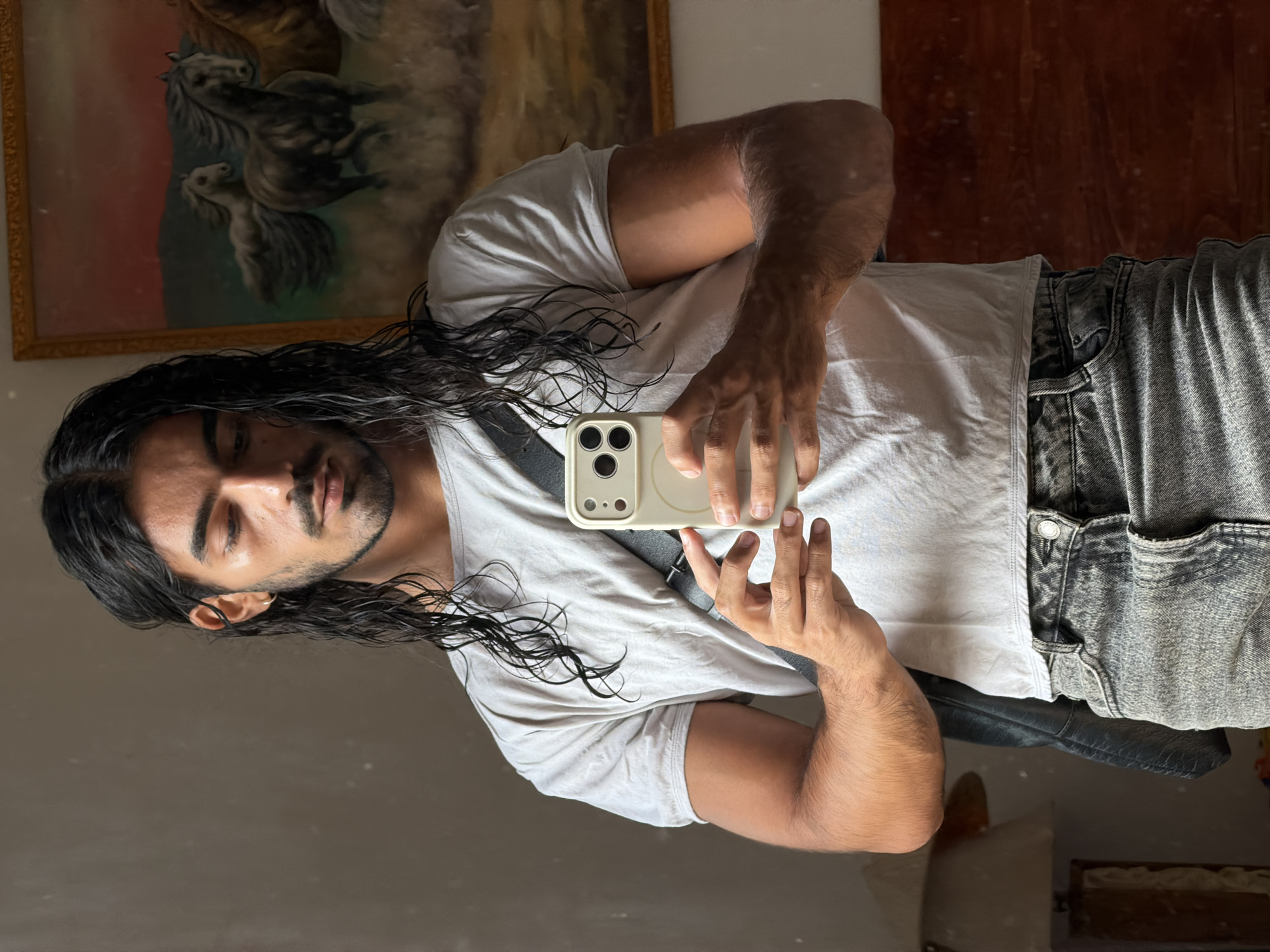 A man with long, dark, wet hair, a light beard, and tan skin is taking a selfie in a mirror. He is wearing a white T-shirt and gray and black jeans. The phone he is holding has a light-colored case. Behind him, there is a framed painting of horses. The reflection in the mirror also shows a dark-colored wall or paneling. The lighting is mixed, with both natural light from a window on the left and artificial light present. The location is North Kuta, Indonesia.