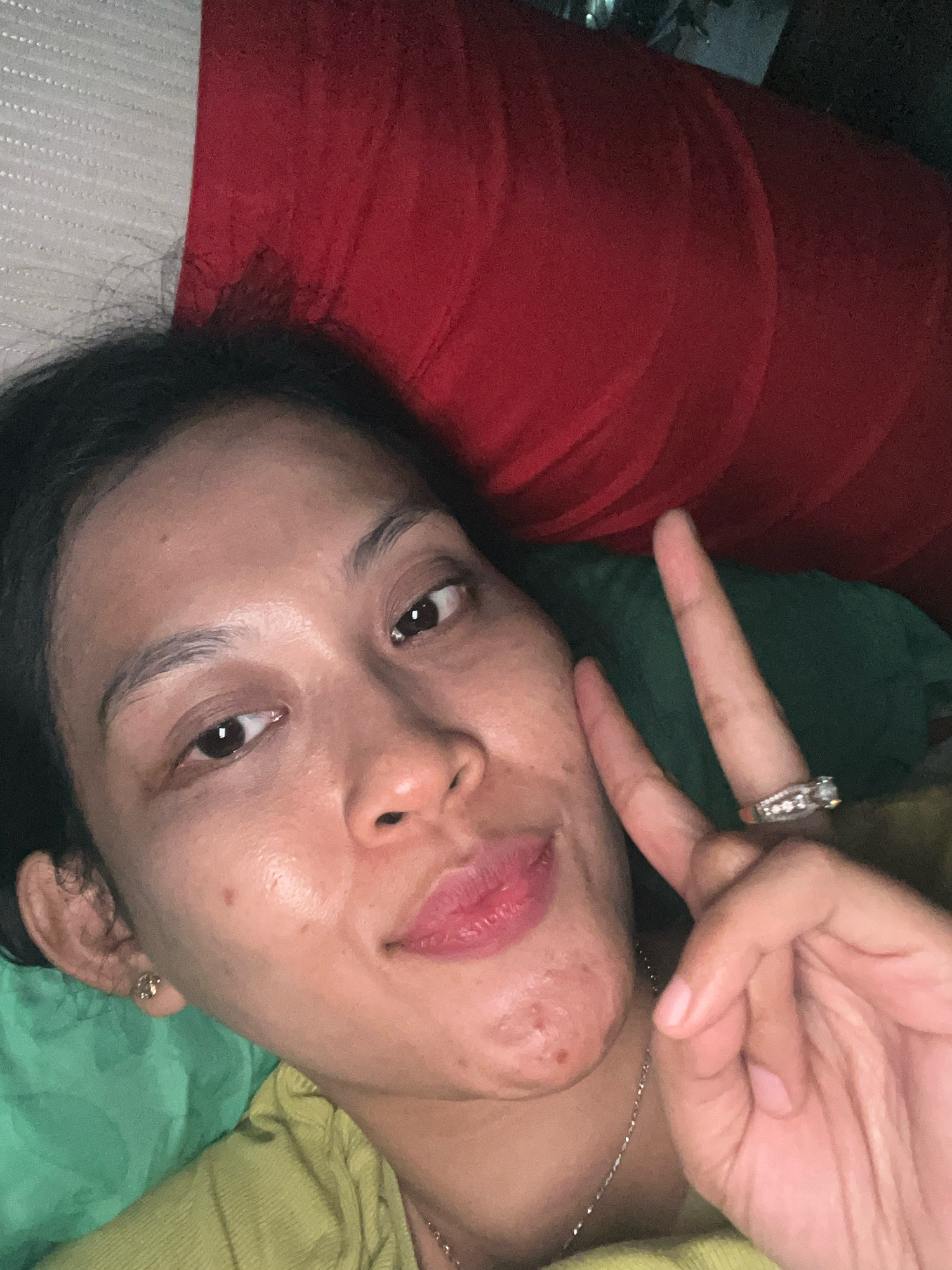 In Kecamatan Pati, Indonesia, a close-up selfie is taken by a woman lying down, resting on red and green pillows. Her face is partially illuminated, revealing a peace sign gesture made with her right hand, adorned with a ring. She is wearing a light green top and a silver necklace. The background consists of textured white and red fabric, suggestive of a home environment.