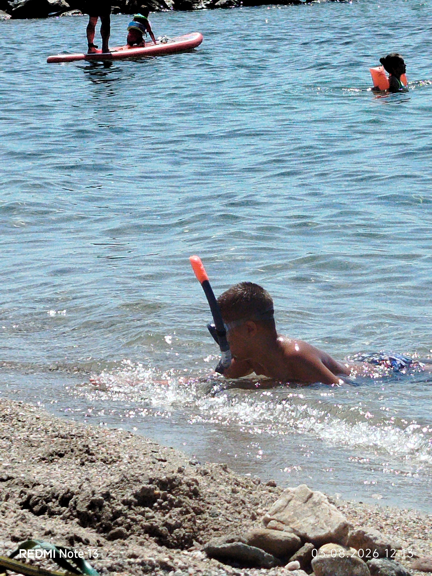 The image depicts a sunny day at a coastal location, likely in Sithonia, Greece, given the presence of clear blue water and a pebble beach. In the foreground, a young person wearing a snorkel and mask is lying in shallow water, with their face submerged. Their body is angled towards the right of the frame, and the water around them sparkles with sunlight. Further in the background, a person stands on a paddleboard, with another individual, possibly a child wearing a life jacket, kneeling on the paddleboard. Another person in the distance is swimming or wading in the water, wearing an orange life jacket. Rocks and a textured beach with pebbles are visible in the bottom portion of the image. The water appears calm with gentle ripples.
