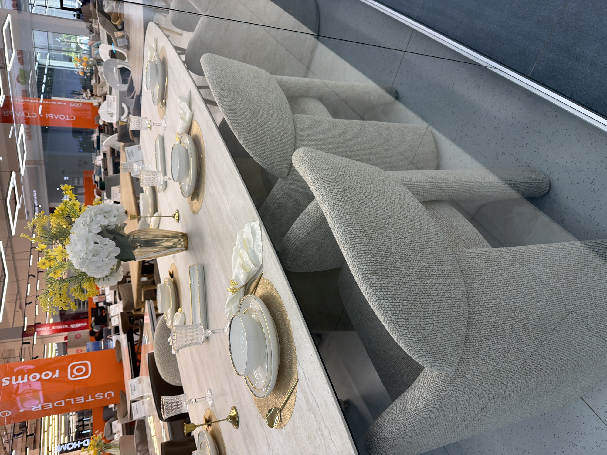 The image captures an indoor scene, likely a furniture showroom, with a polished concrete floor and overhead lighting visible. In the foreground, a rectangular dining table with a light-colored, textured surface is set for a meal. The table is adorned with white plates, bowls, gold cutlery, and white cloth napkins with gold accents. Two clear glass tumblers are also present on the table. To the right of the table, several upholstered chairs with light grey fabric and distinctive curved backs are arranged.
Reflected in a glass partition in the foreground, part of the showroom's interior is visible, including other furniture pieces like tables and chairs, and banners displaying text in Cyrillic and Latin alphabets. One prominent banner is orange and features the text "rooms" and an Instagram logo.
The scene is static and appears to be a staged display rather than a candid moment. No people are actively interacting within the frame. The presence of furniture, displays, and a showroom environment suggests a commercial setting. The text on the banner, "СТОЛЫ СТУЛЬЯ. AZEMANA", which translates to "Tables Chairs. AZEMANA," and the overall aesthetic of the showroom suggest it could be a furniture store located in Astana, Kazakhstan, a city known for its modern architecture and growing retail sector. The presence of international brands and styles is also common in such urban centers.