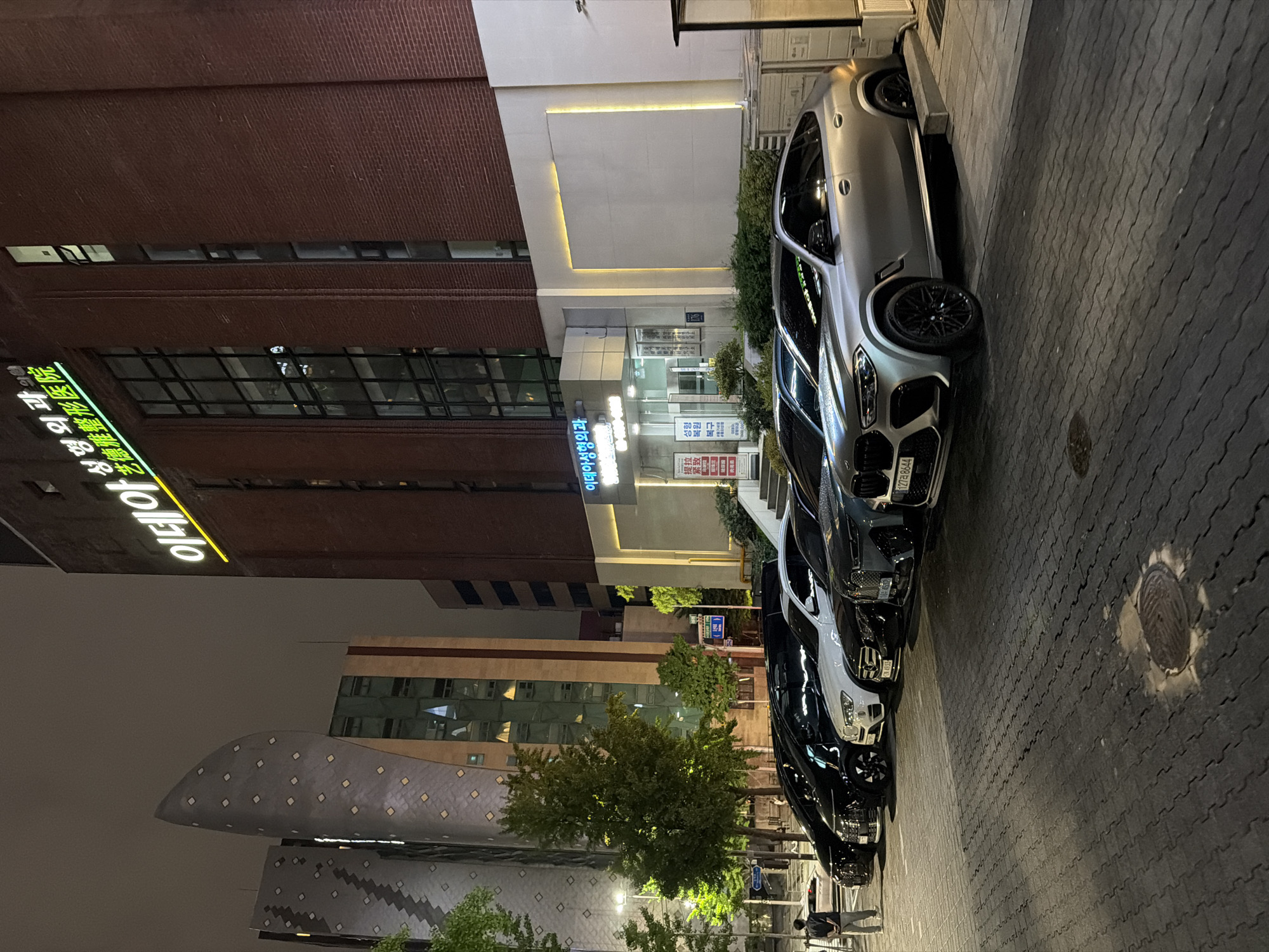 A night-time exterior shot shows a row of parked cars in front of a building in Seoul, South Korea. The building has a red brick facade and illuminated signage in Korean. To the left of the brick building, a modern, curved building with a patterned exterior is visible. The cars are parked on a paved surface, with a person standing in the far left of the frame. The foreground features a manhole cover. The overall scene depicts an urban environment at night, with artificial lighting illuminating the buildings and cars.
