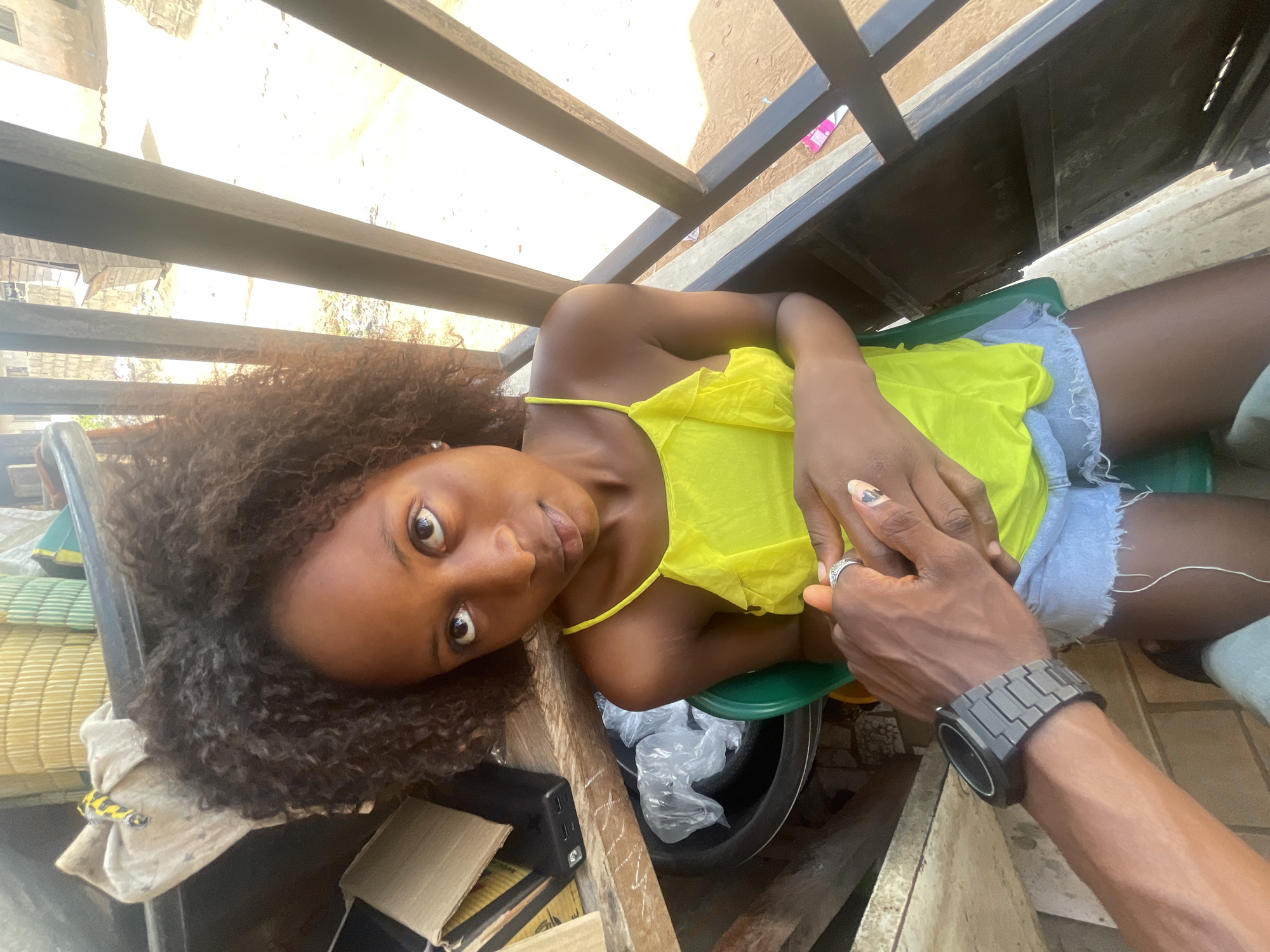 A woman lies prone, her head to the left of the frame, on a surface obscured by items including a black plastic bucket containing plastic bags, a green plastic stool, and cardboard boxes. She wears a yellow top and denim shorts. Her dark curly hair is disheveled around her head. A hand, presumably belonging to a man, grasps her hand. The man is wearing a black watch. Above, a structure of dark metal bars is visible. The location is likely Lokoja, Nigeria.