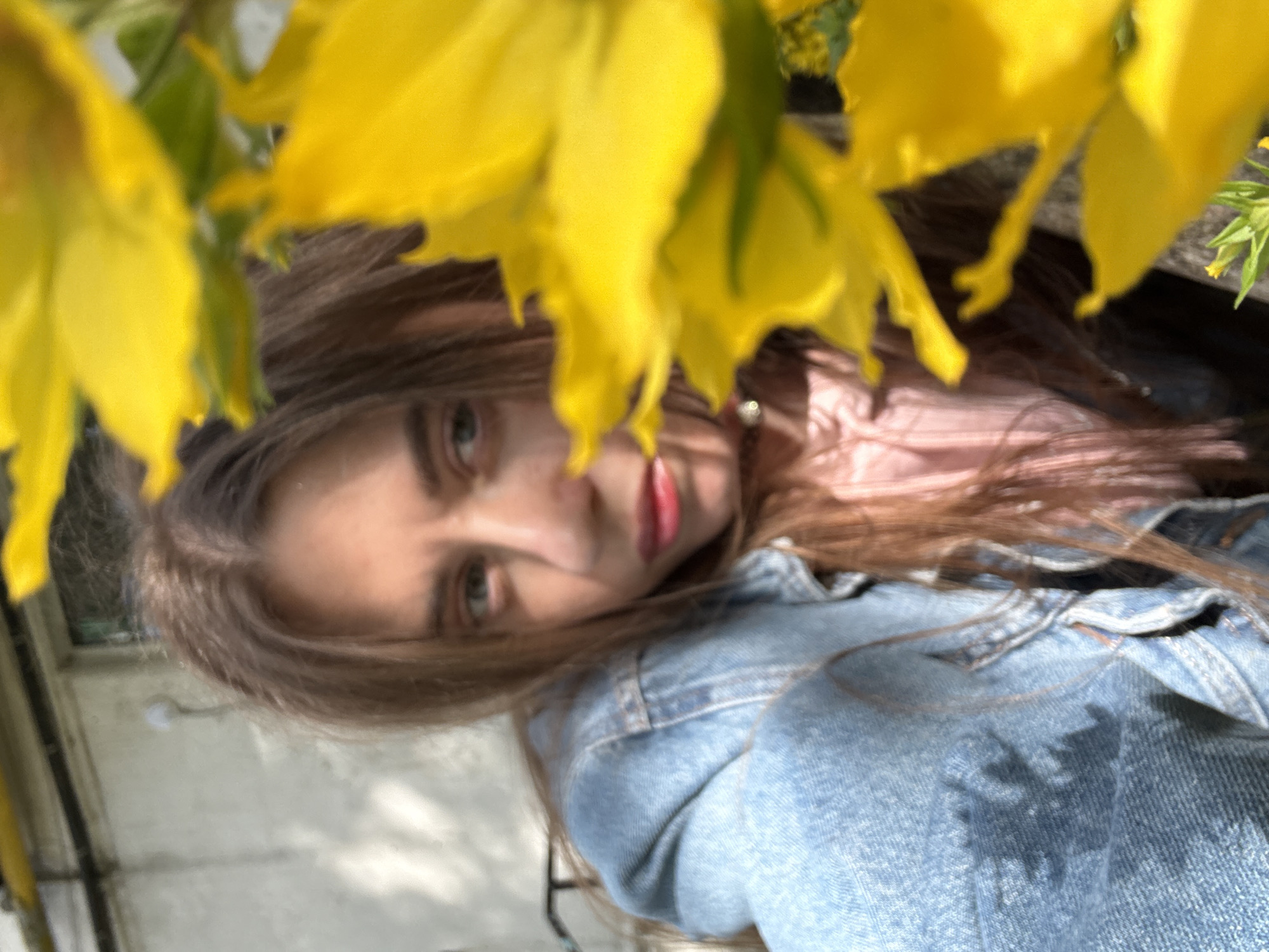 A woman, with shoulder-length brown hair, wears a light blue denim jacket and a pink shirt. She is positioned in the lower half of the frame, looking slightly to the left. Her face is partially obscured by bright yellow flowers in the foreground. The background appears to be a building with a textured, light-colored wall. The scene is outdoors, with sunlight casting shadows. The location is described as Unknown, Unknown.