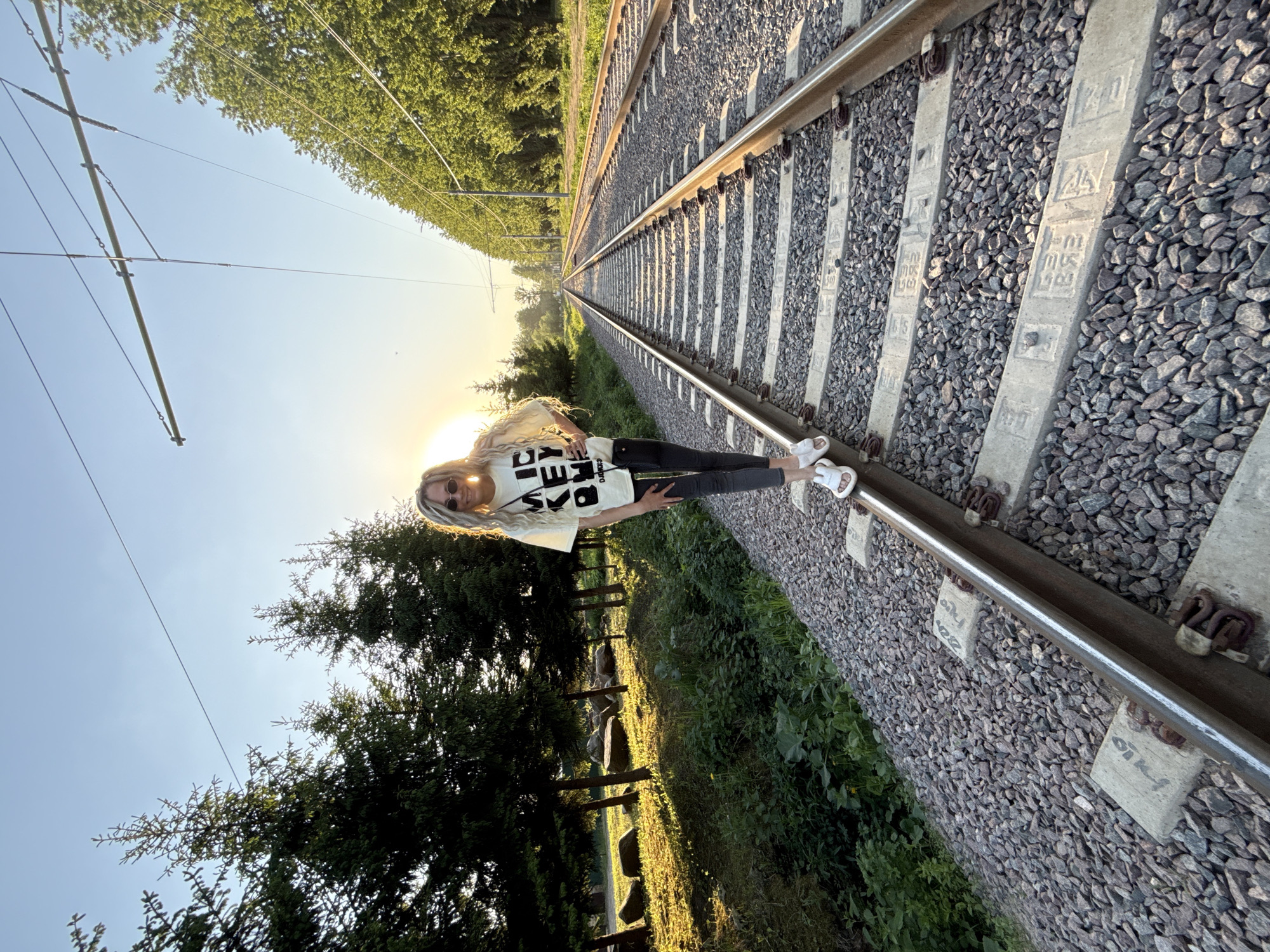 A young woman with long blonde hair, wearing a white t-shirt with black lettering, black pants, and white sandals, is standing on a railway track. She is positioned slightly to the left of the center of the frame. The railway track recedes into the distance, leading towards the setting sun on the horizon. On either side of the tracks, there is green vegetation, including trees and shrubs. Overhead electrical lines run along the tracks. The sky is blue and clear, with the bright sun partially obscured by the woman's head. The railway tracks are made of steel rails and concrete sleepers, with gravel between them. Based on visual cues, the setting is likely near Заволжье, Russia, as the railway appears to be a part of the Gorodets–Zavolzhye railway.