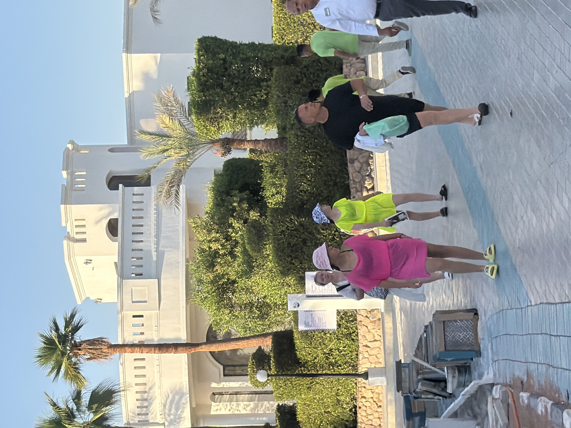 This image captures a sunny outdoor scene likely at a resort in Sharm El Sheikh, Egypt. In the foreground, a group of people are walking along a paved pathway. A man in a black t-shirt and shorts walks towards the viewer, carrying a light green plastic bag and a white cloth. To his left, a woman in a bright pink dress and a pink hat walks away from the viewer, wearing yellow sandals. To the left of the woman in pink, another woman in a white dress and a patterned bag is also walking away. Further in the background, a person in a neon yellow dress is walking. Behind the people, a row of neatly trimmed green bushes forms a backdrop. Beyond the bushes, a large white building with architectural elements reminiscent of a fortress, including battlements and arched openings, stands under a clear blue sky. Two palm trees are visible, one in the mid-ground and another to the left. The pathway appears to be under construction or undergoing maintenance, with some materials and equipment like buckets and wooden crates visible on the left side of the image. The overall impression is of a leisurely, warm-weather vacation environment.