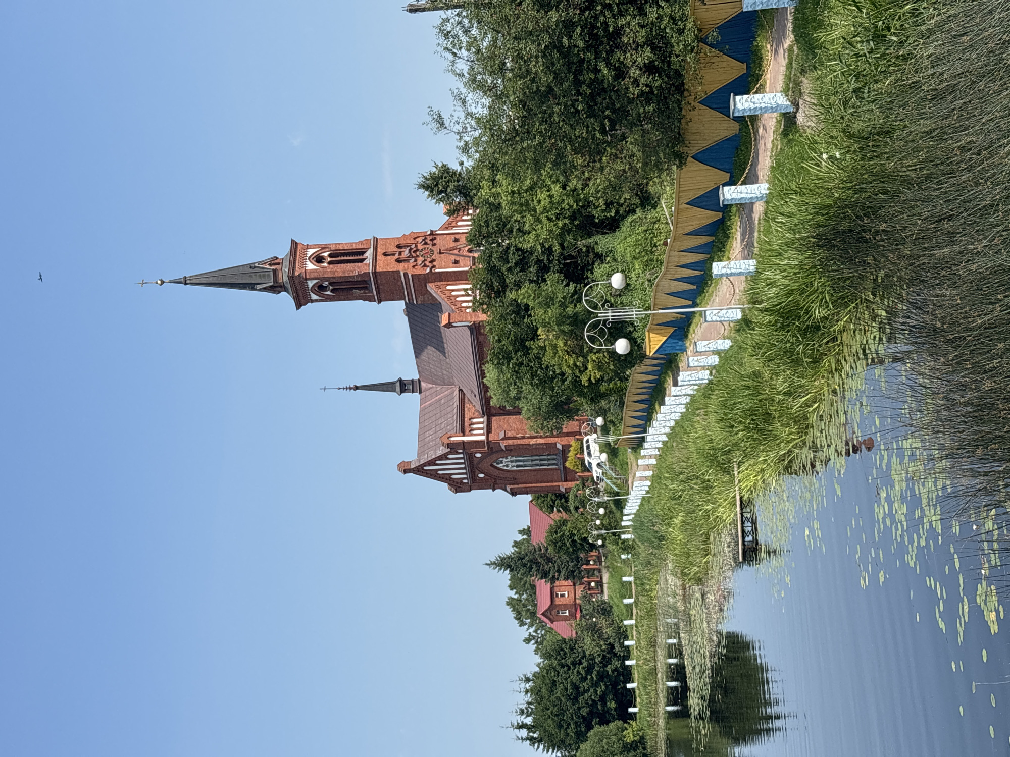 In Pastavy, Belarus, a large, red brick church with a tall, dark spire stands prominently on a grassy hill. The church features Gothic architectural elements, including pointed arches and white decorative trim. Adjacent to the church, a smaller red-roofed building with white trim is visible. A white car is parked near the church entrance.
In the foreground, a body of water, likely a lake or river, is present. The water's surface is dotted with lily pads and reflections of the surrounding greenery and sky. Tall reeds and grass grow along the water's edge.
A pathway lined with decorative white posts and a yellow and blue zigzag fence leads away from the water and up towards the church. Several white lampposts with spherical lights are positioned along this pathway. The sky above is clear and blue. A small, dark object, possibly an aircraft, is visible in the upper right portion of the sky. The scene depicts a serene, picturesque view of a religious institution and its natural surroundings.