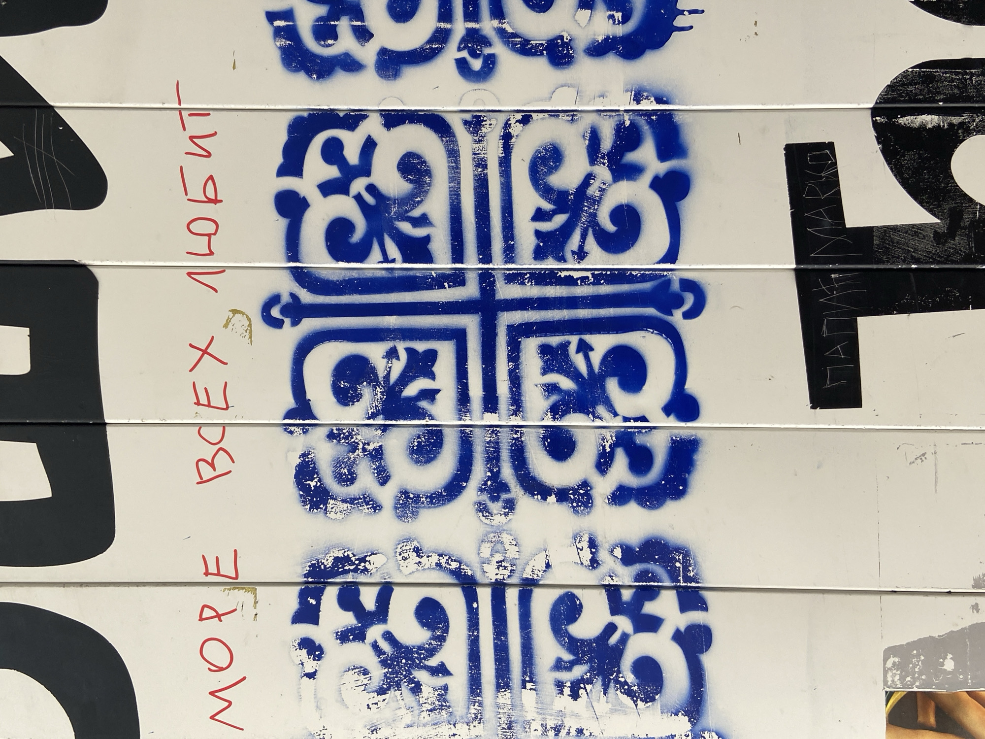 This media file is a close-up shot of a white surface with blue stencil art and red handwritten text. The stencil art features intricate, repeating patterns with swirling motifs and geometric shapes, primarily in blue. The white surface appears to be made of horizontally layered panels, suggested by thin lines running across the image. The texture of the surface shows signs of wear and tear, with paint chips and scuff marks visible, indicating it might be an outdoor surface or an old wall.
Scattered across the image are pieces of black graphic elements, some appearing to be letters or parts of larger designs, partially overlapping the blue stencil art. One prominent black shape on the right side resembles a stylized capital letter, possibly part of a larger advertisement or artwork.
Red handwritten text in Cyrillic script is visible on the left side of the image, oriented vertically and upside down relative to the blue stencil art. The legible parts of the text include "ВСЕХ ЛЮБИТ" and "MORE". The handwriting is rough and appears to be done with a marker or crayon.
On the right side of the image, some faint black lettering is scrawled, appearing to be "МАРКО" in Cyrillic. There are also some small, torn pieces of what look like paper or photographs at the bottom right, revealing a sliver of human skin and fabric, possibly from a magazine or poster.
The overall impression is one of urban decay or street art, where multiple layers of artistic expression and graffiti have been applied to a weathered surface. The lighting is even, suggesting it might be daytime, but no specific time or weather cues are apparent. The setting is likely an exterior urban environment.