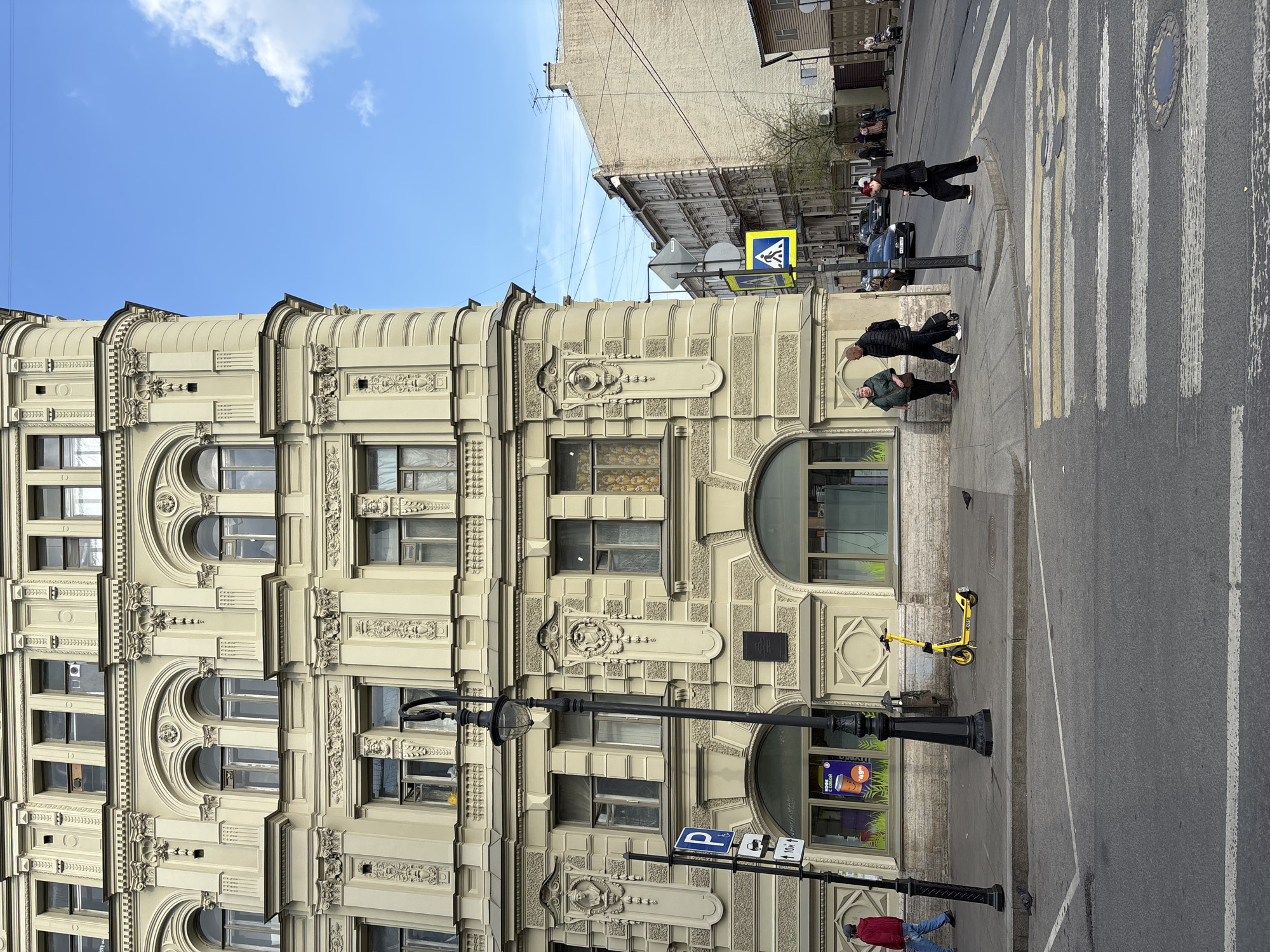 The image depicts a multi-story building with ornate architectural details, located in Saint Petersburg, Russia. The building is beige, with decorative window frames, moldings, and architectural embellishments. A street scene is in front of the building, with a black lamppost, street signs, and a yellow scooter. People are walking on the sidewalk and crosswalk. The sky is blue with a few clouds. A neighboring building is partially visible behind the primary building.