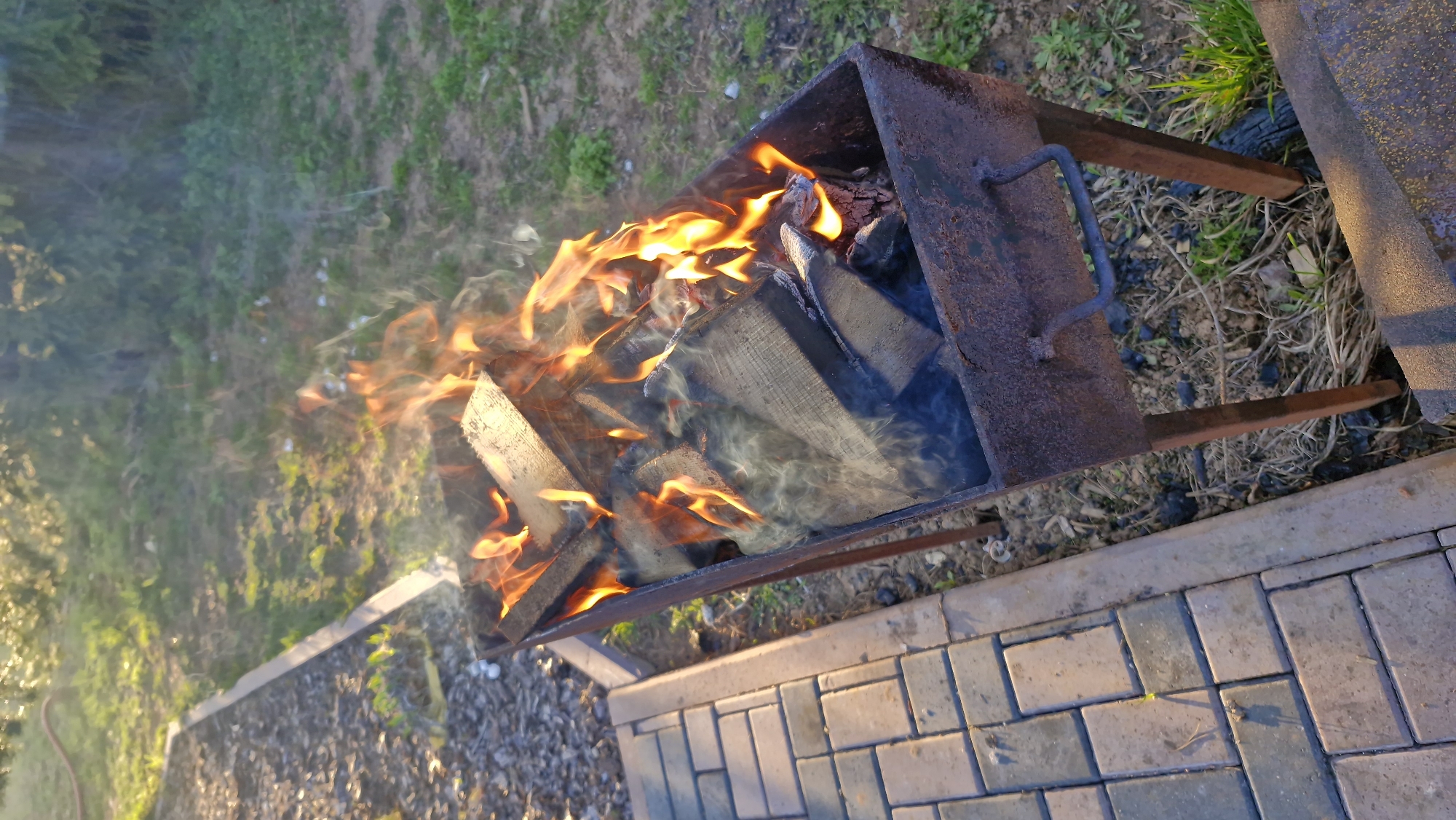 A rusty, metal brazier sits on a brick-paved area. Flames engulf split logs of wood inside the brazier, creating smoke that drifts to the left. The area is adjacent to a patch of grass and foliage. The photograph was likely taken at Otchor, Russia.