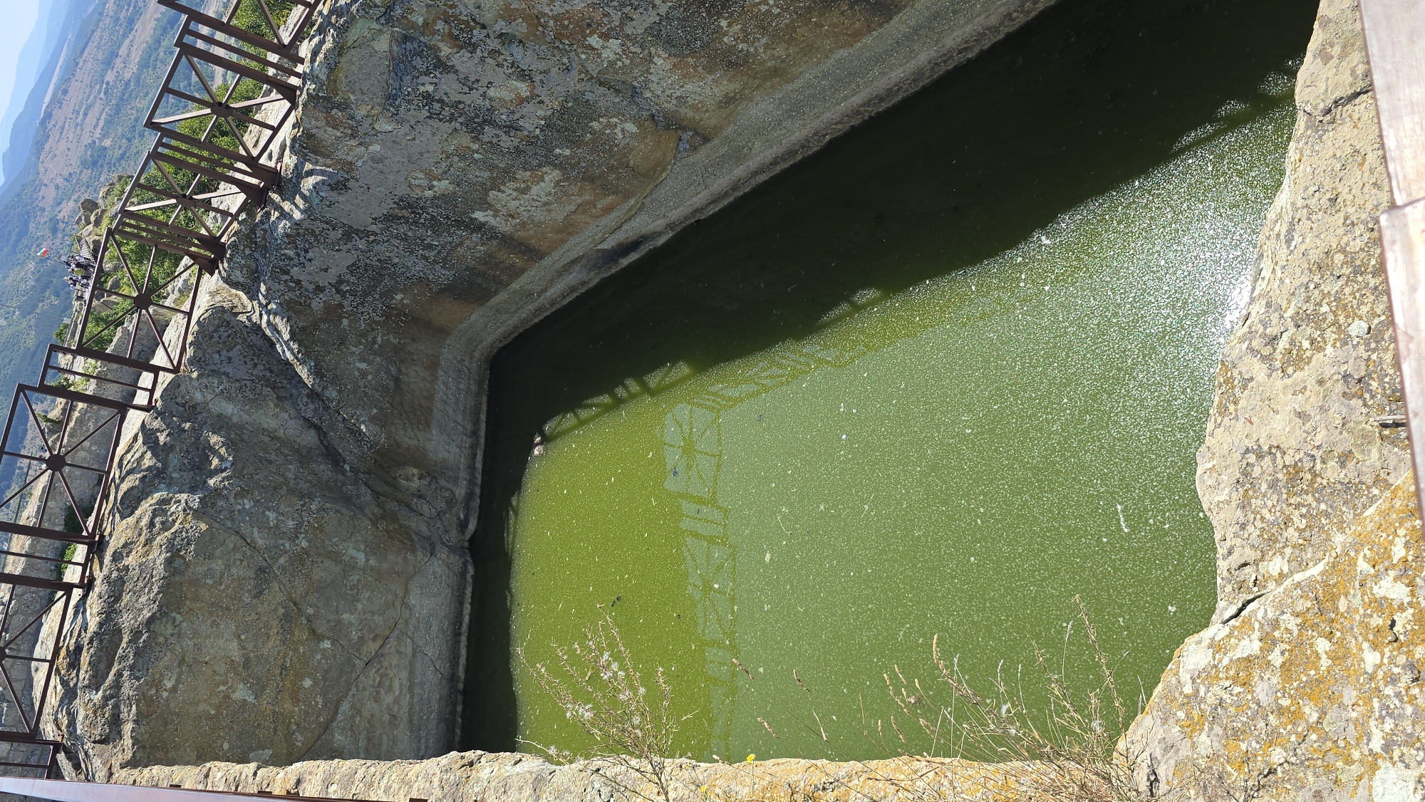 The image shows an ancient Thracian rock sanctuary located in the city of Gorna Krepost, Bulgaria. The sanctuary features a large, rectangular rock-cut pool filled with murky green water. The pool's edges are made of rough, weathered rock, and some areas appear to be reinforced with concrete or mortar. A metal railing with a lattice design is visible along the top edge of the pool, providing safety for visitors. Beyond the railing, the landscape is hilly and mountainous, with sparse vegetation and distant hills under a clear blue sky. Several people are standing near the railing in the background, observing the site. The overall scene depicts a historical and archaeological site integrated into a natural landscape.