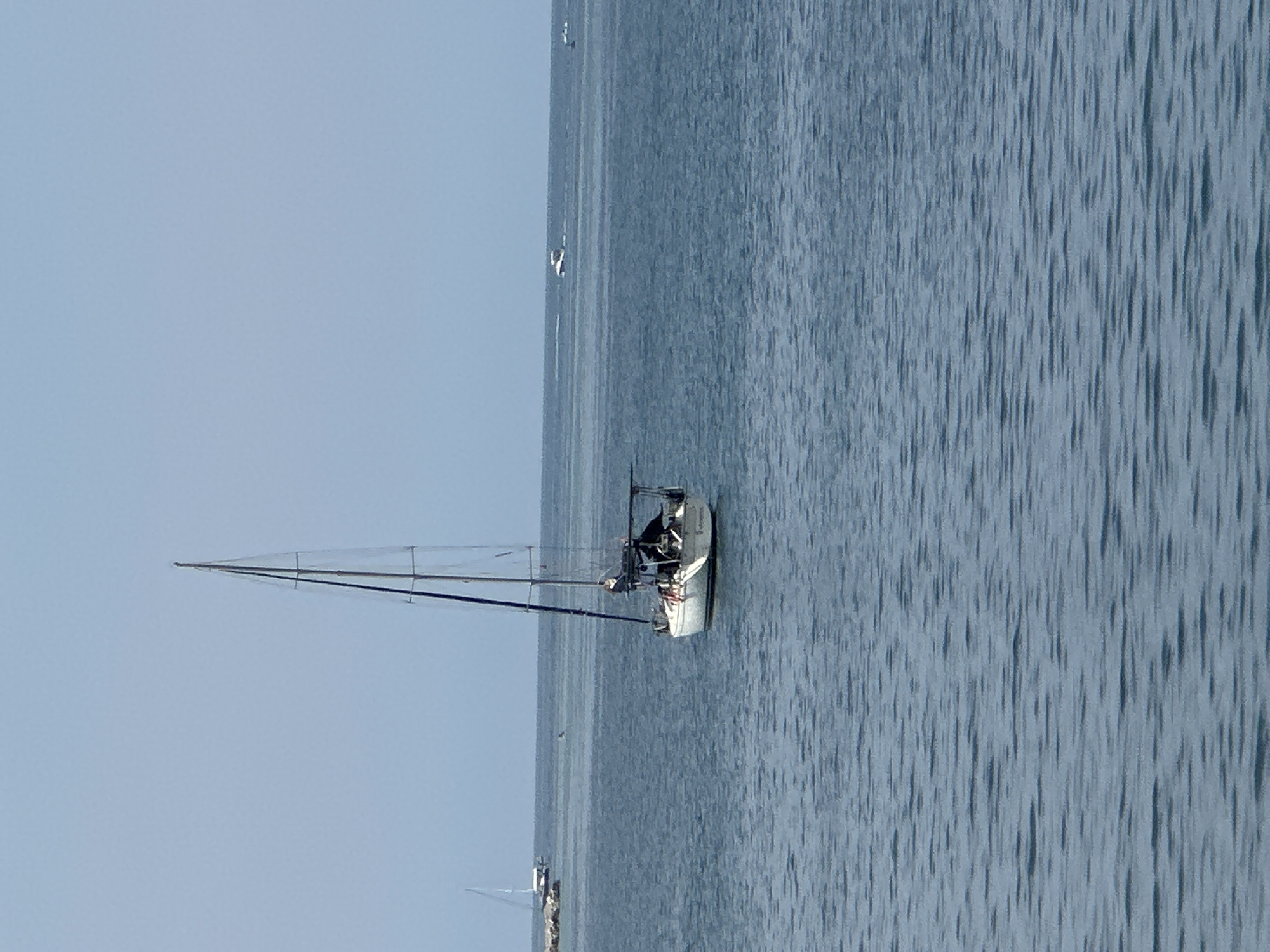 A sailboat is anchored in calm blue water. The sailboat is white with a dark blue sail and a dark canopy over the stern. There are a few people on the sailboat, one of whom appears to be steering. In the distance, there are other boats, including a white motorboat and another sailboat. The sky is clear and pale blue. The scene appears to be in Budva, Montenegro, based on visual cues from similar locations.