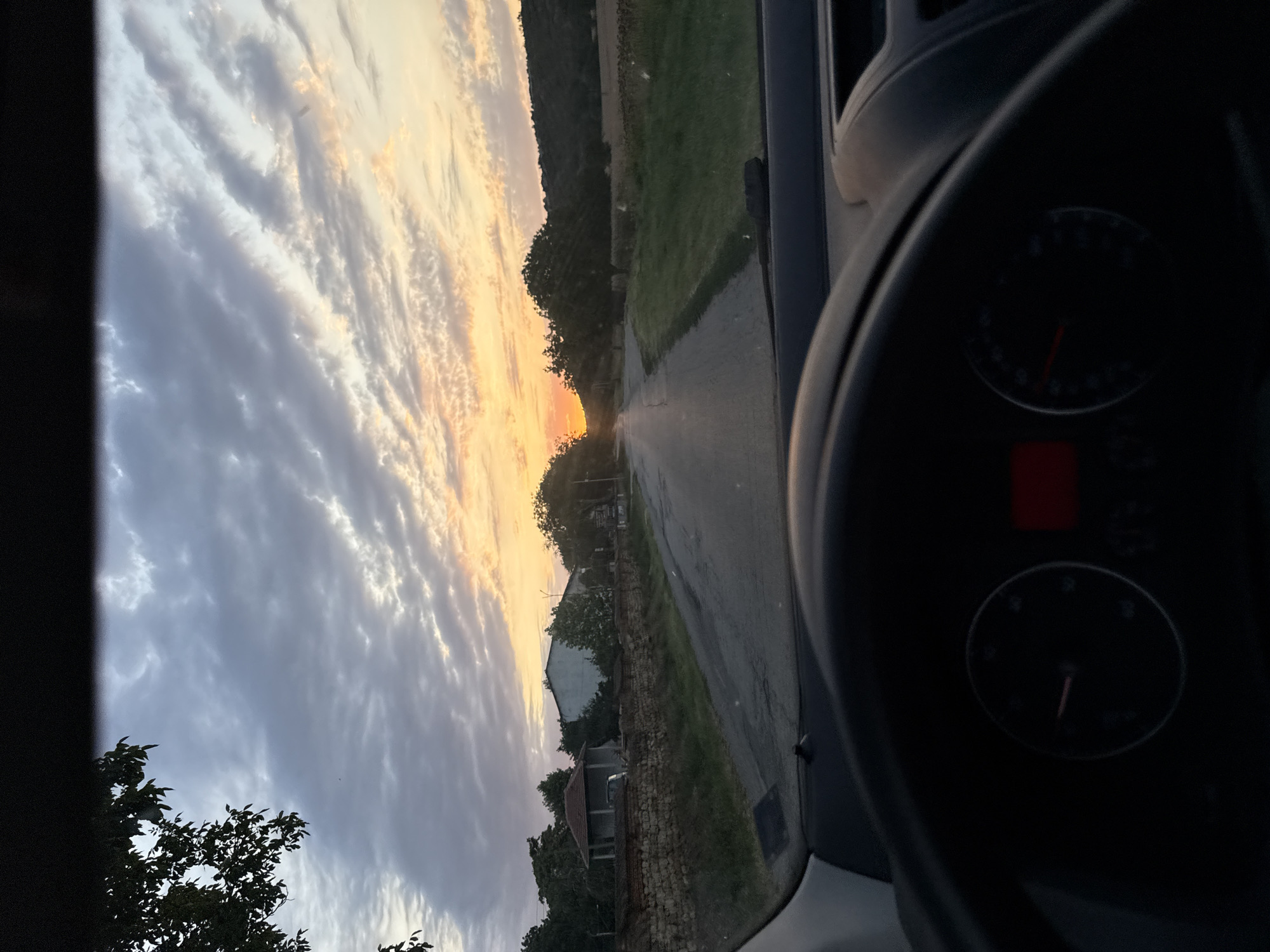 The image is taken from the interior of a vehicle, looking out through the windshield at a rural road at sunset. The sky is filled with dramatic, striated clouds catching the warm hues of the setting sun. A tree branch hangs into the frame from the upper left. The road stretches into the distance, leading towards the horizon where the sun is setting behind a line of trees and rolling hills.
On the left side of the road, there is a low, stone wall, beyond which are some buildings and trees. One building appears to be a small, open-sided structure with a red-tiled roof, and next to it is a larger, white building. Further down the road, on the right side, there is a field with some dry vegetation and then a wooded hillside. The foreground of the image is dominated by the dark silhouette of the car's dashboard and steering wheel, with a partially visible speedometer showing a speed reading. The overall impression is of a quiet, picturesque drive through the countryside as day transitions into evening.
This scene is located in Slavyani, Bulgaria.