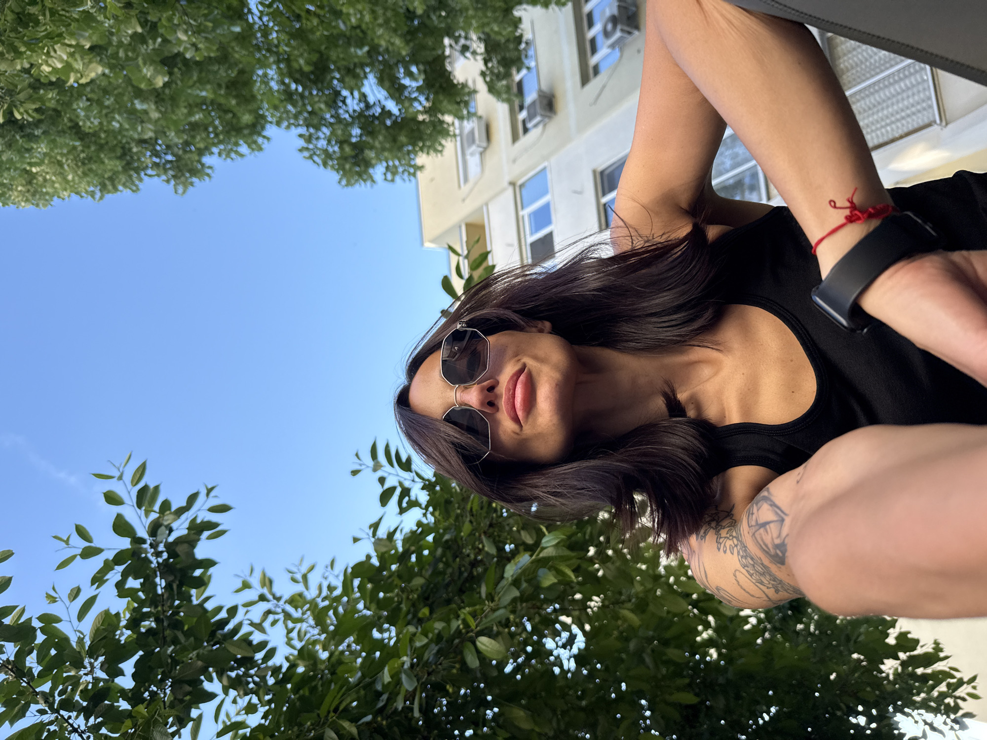 The image is a low-angle selfie of a woman with dark hair and sunglasses. She is wearing a black tank top, and her arms are crossed. There is a tattoo visible on her arm, and she is wearing a smartwatch and a red string bracelet on her wrist. The background includes green trees and a building. Based on the architectural style of the building, it could be located in Pleven, Bulgaria. The sky is blue and clear.