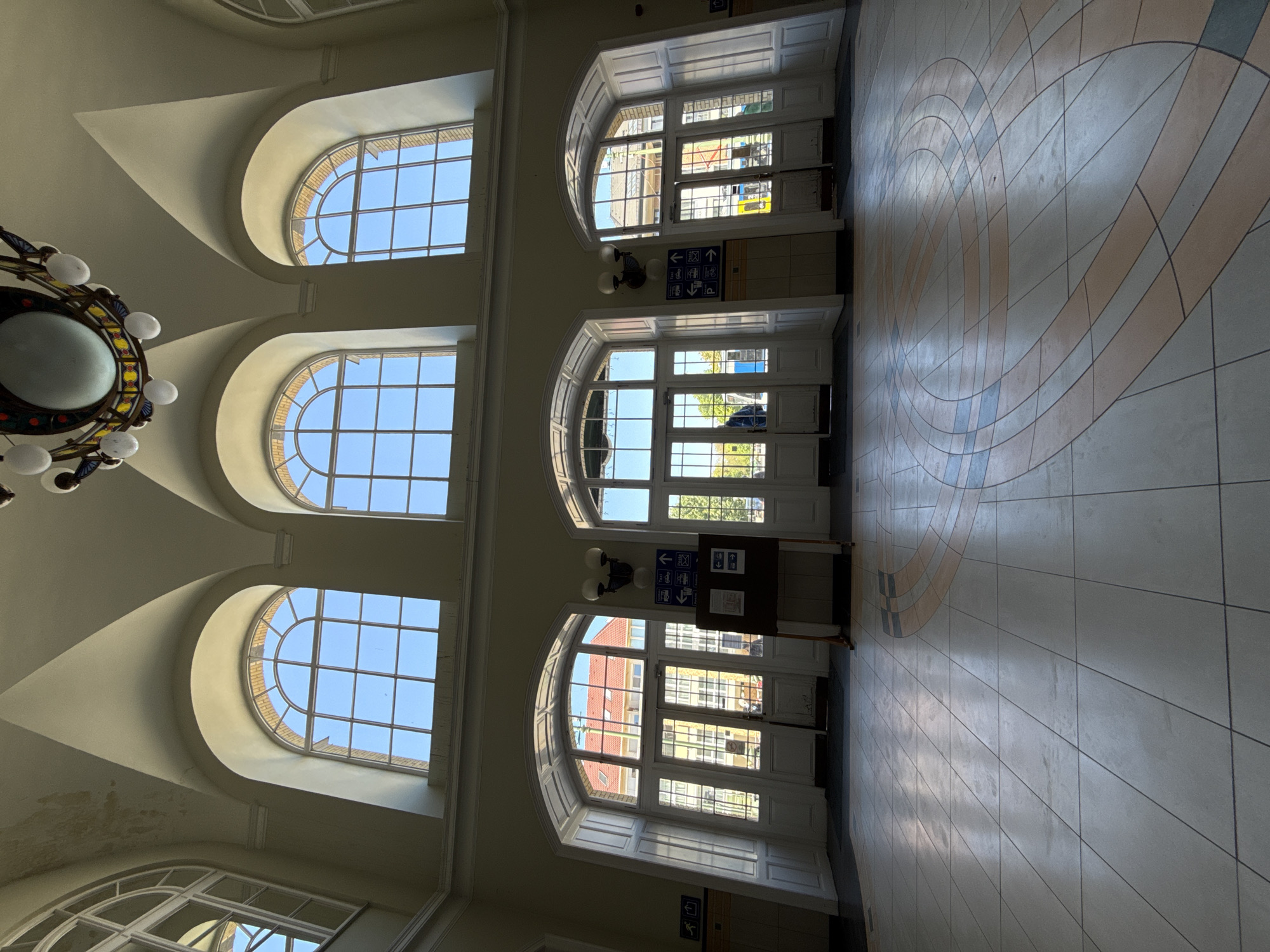 The image captures the interior of a large, grand public hall or station, located in Szeged, Hungary. The scene is dominated by classical architectural features, including high ceilings with coved arches, large arched windows, and equally grand arched doorways.
On the left side of the frame, a series of three tall, arched windows allow abundant natural light to flood the space. Through these windows, a clear blue sky is visible, along with glimpses of a brick building exterior. From the ceiling on the far left, a decorative chandelier with multiple bulbs and a central globe hangs.
On the right side, two large arched openings serve as main entrances or exits. These feature paneled white doors with glass sections, revealing views of distant buildings and trees outside. Between these two lower arched doorways, a dark brown information stand is positioned, featuring several blue directional signs with white pictograms and arrows. These signs clearly indicate directions for "train" (left arrow), "bus" (right arrow), "parking" (down arrow), and an icon for "lodging/hotel" (left arrow). A smaller blue sign with a right-pointing arrow and a stylized human figure is also visible near the floor on the lower left.
The floor is covered with light-colored tiles, featuring a prominent, large circular geometric pattern in shades of cream, beige, and light blue/grey. The bright natural light streaming in suggests it is daytime, likely morning or early afternoon, creating distinct areas of sunlight and shadow across the floor. No people are visible, lending a quiet and spacious feel to the scene. The overall impression is one of historical elegance combined with functional public utility.