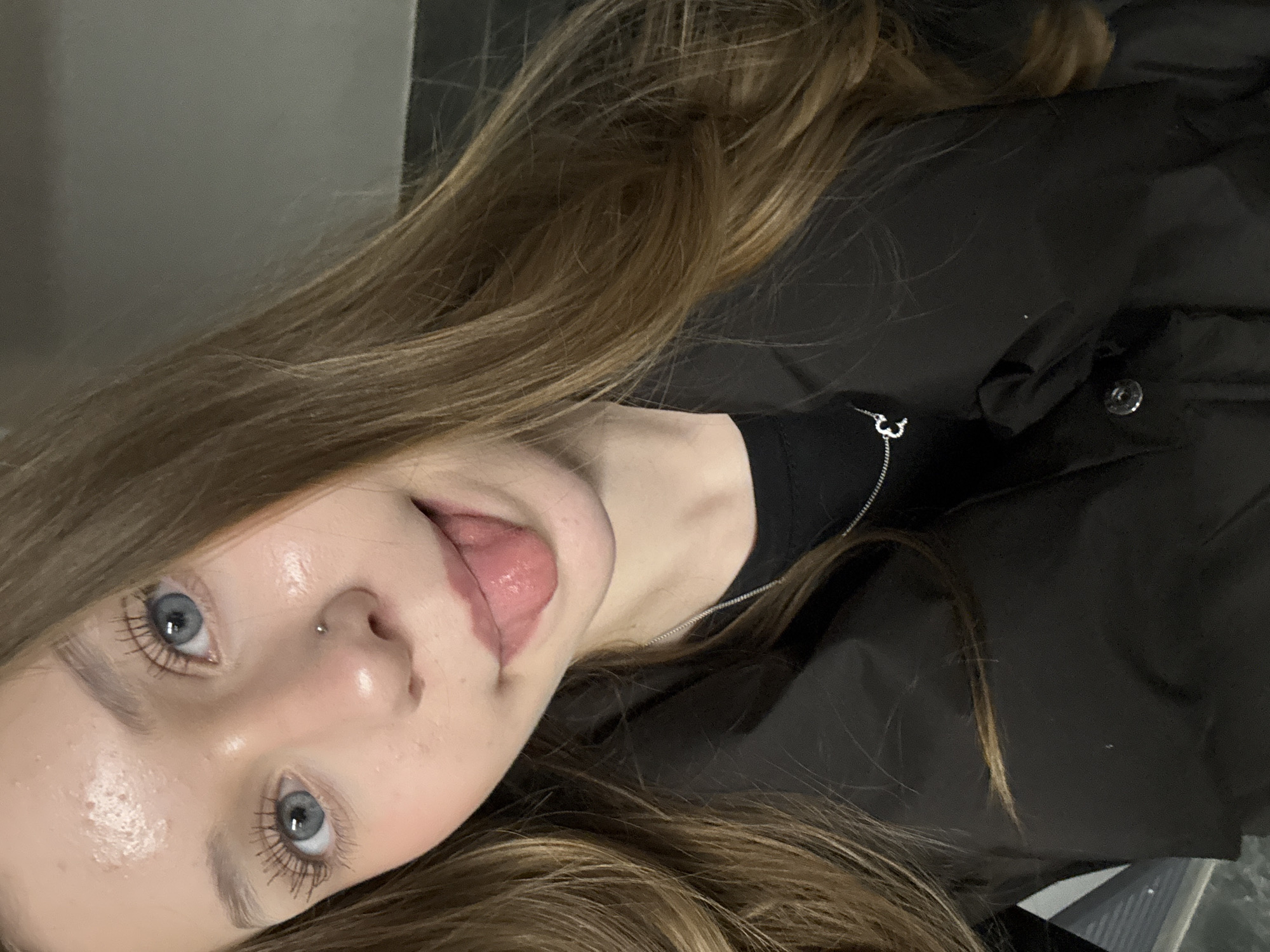 A young woman with light brown hair and blue eyes is looking at the camera, her tongue slightly out. She has a nose piercing. She is wearing a black shirt and a black jacket. The background is gray and appears to be indoors. There is no information to suggest this image was taken in Yekaterinburg, Russia.