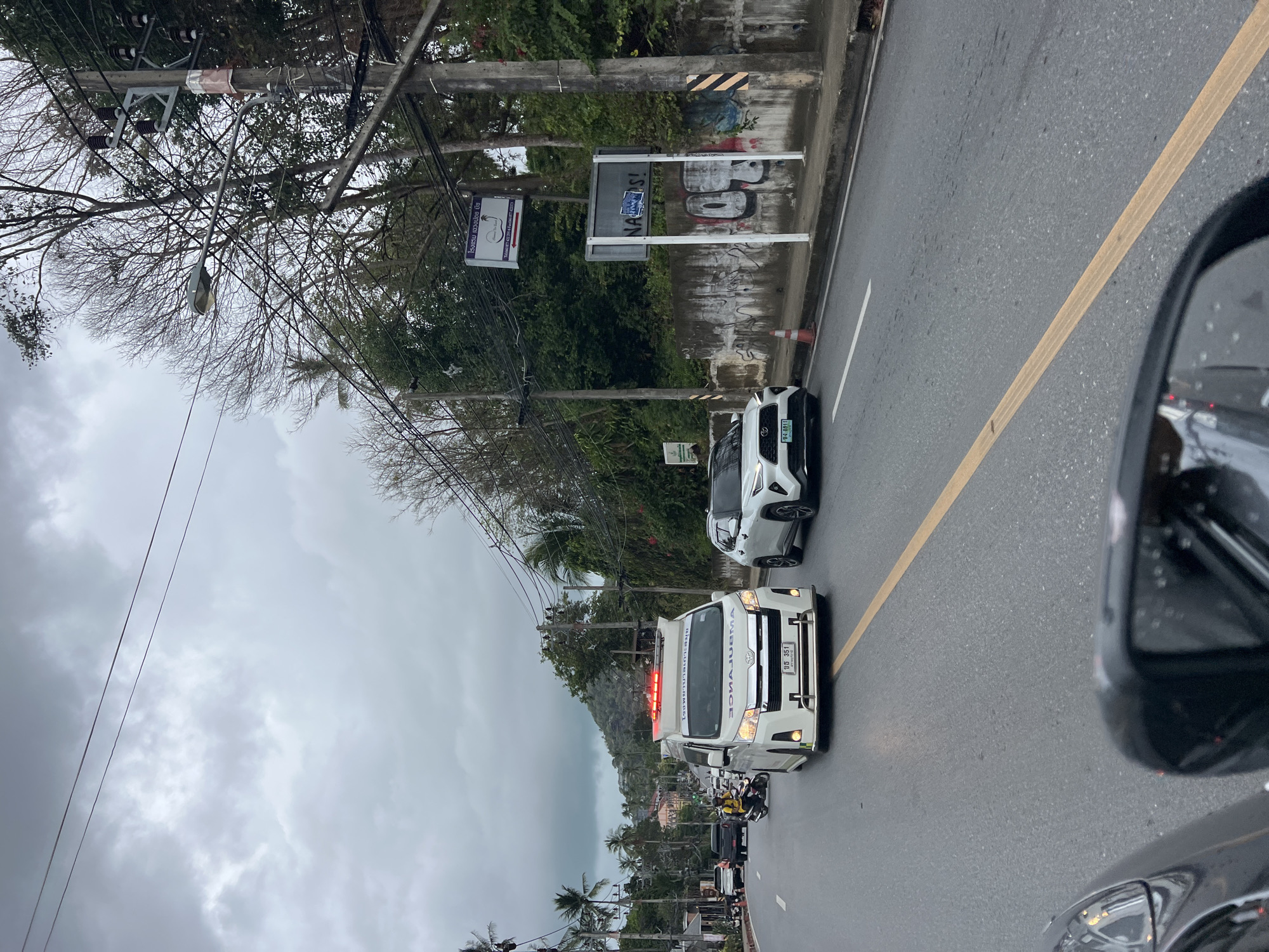 The image shows a street scene in Baan Chaweng Noi, Thailand, on an overcast day. An ambulance with flashing red lights is positioned in the middle of the road, followed by a white SUV. Traffic is present in both directions. Power lines run along the side of the road, and a partially obscured sign is visible on a concrete wall. A rearview mirror of another vehicle is visible in the foreground. Palm trees are visible in the distance.