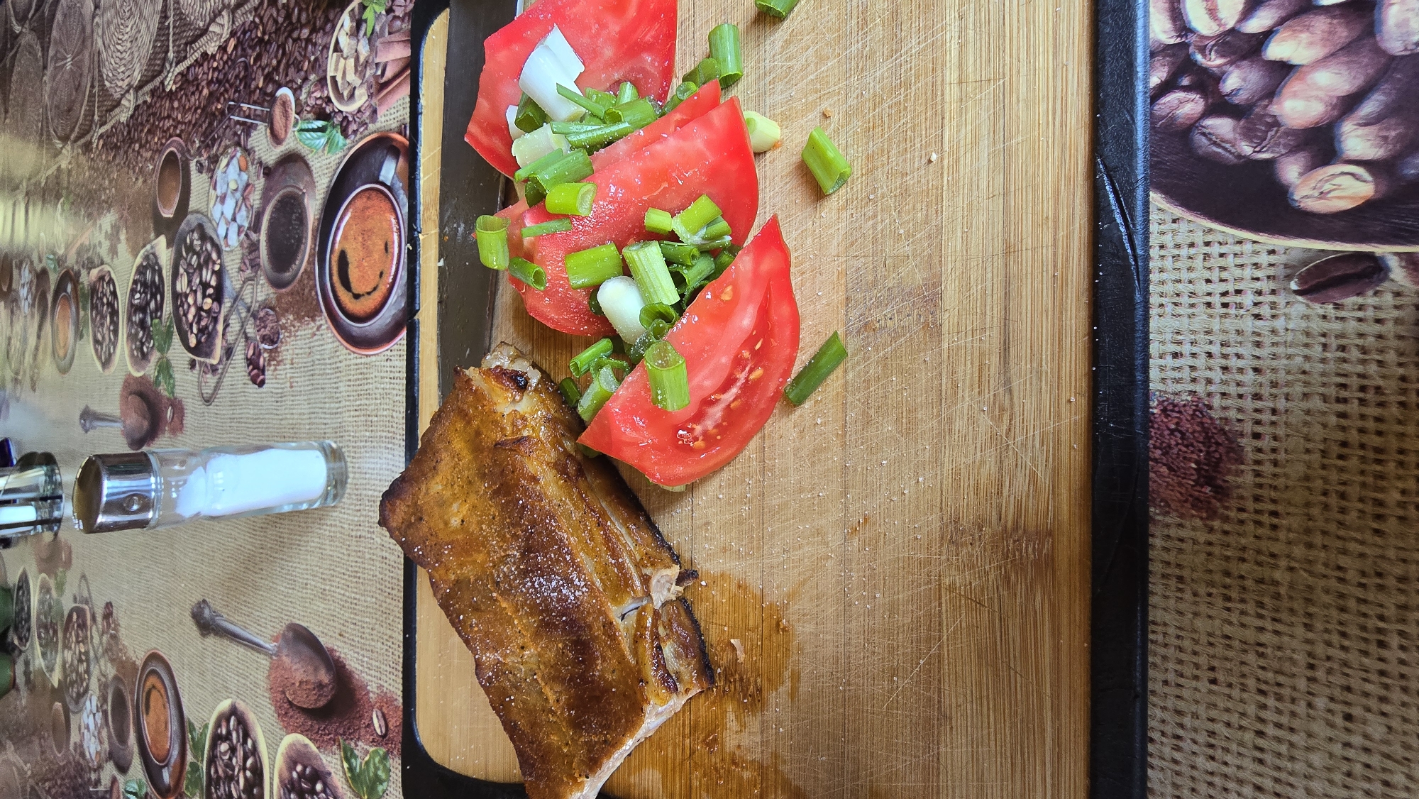 This image captures a meal served on a wooden cutting board, situated on a patterned tablecloth, within a domestic setting in Gorni Bogrov, Bulgaria. The primary subject is a substantial piece of cooked meat, likely a cut of ribs or belly, positioned on the left side of the cutting board. It exhibits a crispy, golden-brown exterior with visible layers of fat and is lightly seasoned with coarse white salt crystals. Adjacent to the meat, on the right side of the board, are multiple thick slices of red tomato, approximately three to four, topped with chopped green scallions and small white fragments, possibly white onion or garlic. Fine salt crystals are also visible on the vegetables.
The cutting board itself is light brown, rectangular, and shows significant signs of use, including numerous parallel and intersecting knife marks and scratches across its surface. In the upper-middle background, a clear glass salt shaker with a metallic silver-colored top stands upright, containing white granular salt. The cutting board rests upon a textured tablecloth patterned with various coffee-related motifs, including coffee beans, ground coffee, cups of brewed coffee, and serving spoons. The tablecloth's base color is a light beige or natural fabric tone, suggesting a woven material like burlap, with the coffee imagery rendered in shades of brown and dark earth tones. The scene is illuminated by an overhead or ambient light source, casting soft shadows.