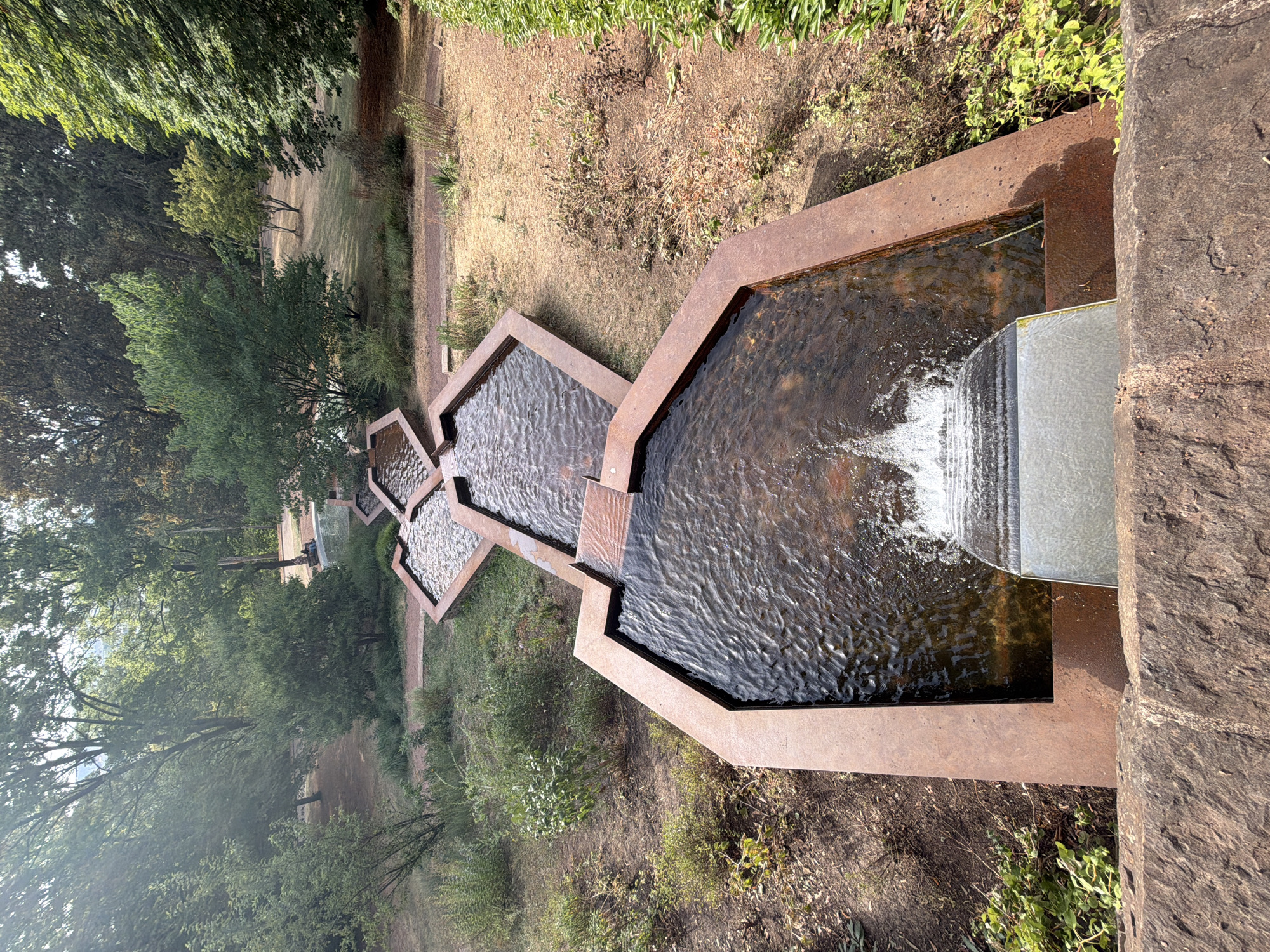 A tiered fountain with rust-colored concrete basins flows with water, cascading from one level to the next. The water appears clear and is gently rippling. At the lowest level, a modern metal spillway directs the water into an unseen reservoir, creating a small waterfall effect. Surrounding the fountain is a naturalistic setting with dry, brown earth and patches of green grass and bushes. Tall, leafy trees with vibrant green foliage flank the scene, suggesting a park or garden environment. Sunlight filters through the trees, casting dappled shadows on the ground. The overall impression is one of a tranquil outdoor space, possibly a park in Esch-sur-Alzette, Luxembourg, given the location context. The time of day appears to be daytime, with bright sunlight. No people or text are visible in the image.