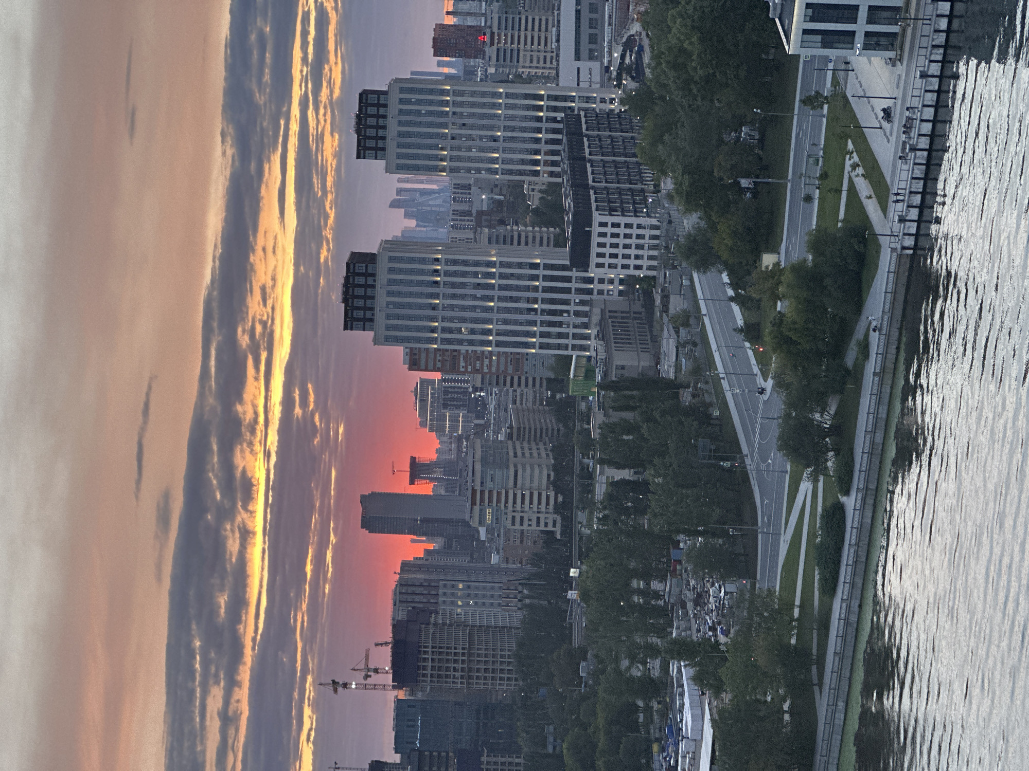 The image depicts a sunset over Moscow, Russia. In the foreground, a body of water with ripples is visible, with a pier or embankment to the right. Further back, there is a park area with trees, pathways, and seating. Across the park, a road with traffic and a few pedestrians can be seen.
The middle ground of the image is dominated by a cityscape with various buildings, including several modern, tall skyscrapers. Two prominent, white, rectangular skyscrapers with dark tops stand side-by-side, illuminated by external lights. In the distance, more skyscrapers are visible, with a striking bright red sunset illuminating the sky behind them. Construction cranes are also present, indicating ongoing development in the city. The sky is filled with clouds tinged with orange, pink, and gray hues from the sunset.