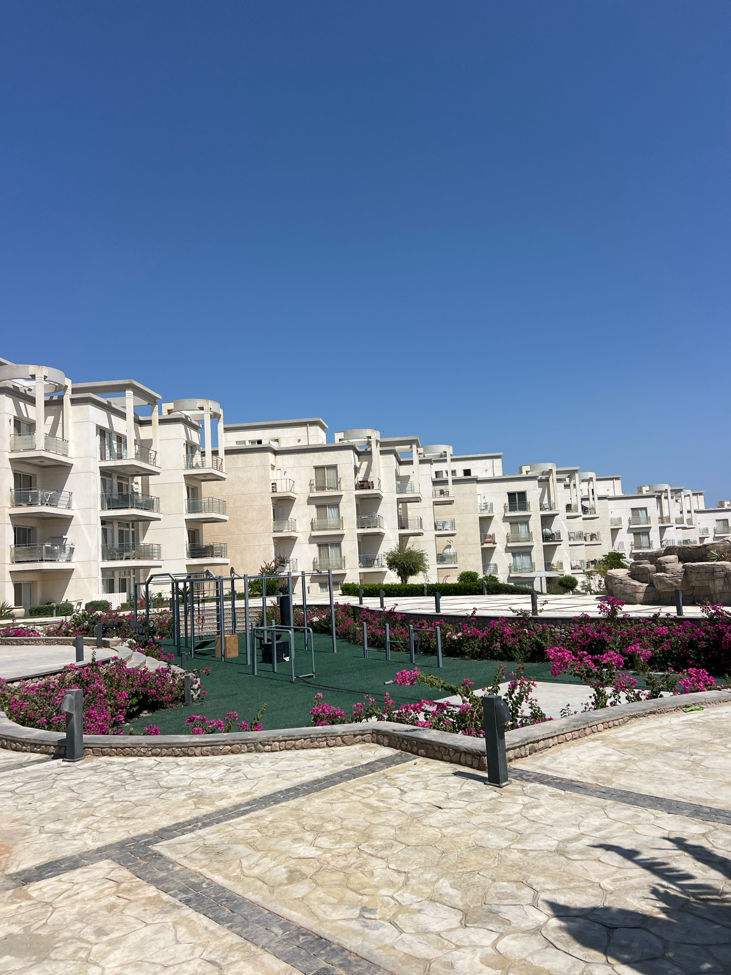 This is a photograph of the exterior of a resort or residential complex in Sharm El Sheikh, Egypt, under a clear blue sky. In the foreground, there is a paved area with a textured, light-colored stone pattern, bordered by a low stone wall. Beyond the paving, there is a circular area with a low stone wall and vibrant pink bougainvillea flowers. Within this area, there is a green turf surface with outdoor exercise equipment, including pull-up bars and other metal structures.
In the background, there is a series of multi-story buildings with a modern architectural style, characterized by white facades, balconies with glass railings, and rounded decorative elements on the upper levels. The buildings are arranged in a stepped or staggered formation. To the far right of the image, there are artificial rock formations. The overall scene depicts a well-maintained outdoor recreational area within a resort setting. There are no people visible in the image.